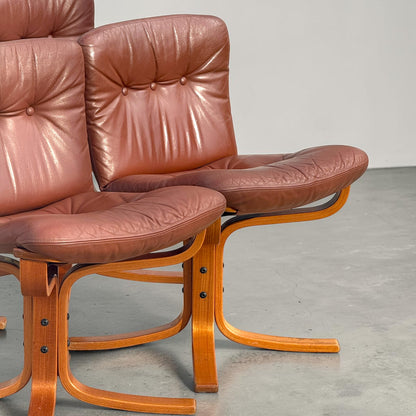 A close-up of two Rare Vintage Joe Rufenacht Lounge Chairs from a set of 4, featuring Howe brown leather and curved wood frames in Scandinavian style, placed on a light gray concrete floor against a plain wall.