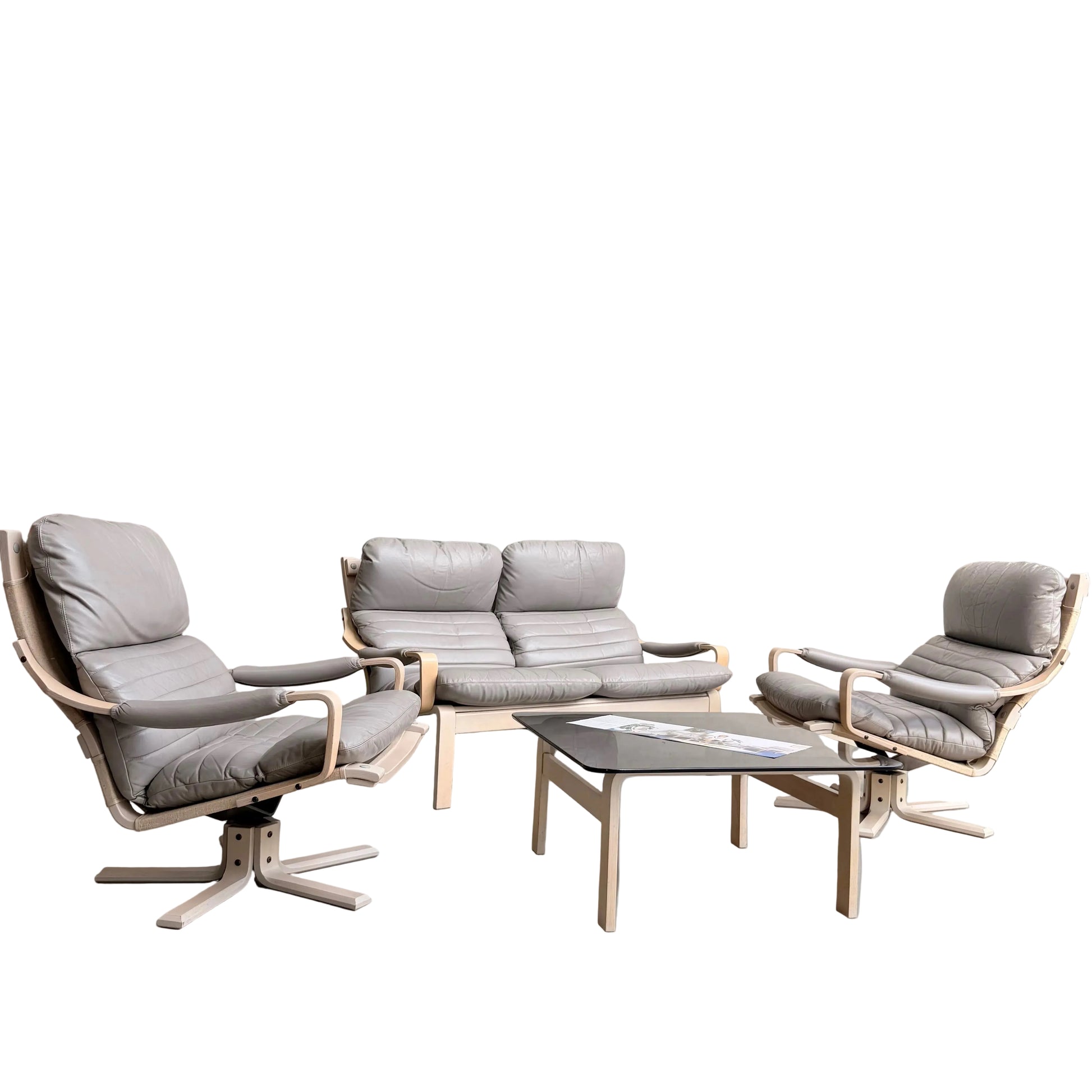 A minimalist room with pale gray walls features the Tessa Ferrara Lounge Suite – Complete 5-Piece Set (Revive) in light wood and gray leather. Sunlight streams in from the right, highlighting the sofa, two armchairs, and rectangular coffee table with magazines.