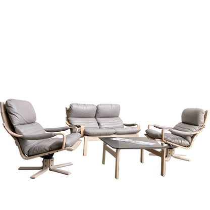 A minimalist room with pale gray walls features the Tessa Ferrara Lounge Suite – Complete 5-Piece Set (Revive) in light wood and gray leather. Sunlight streams in from the right, highlighting the sofa, two armchairs, and rectangular coffee table with magazines.