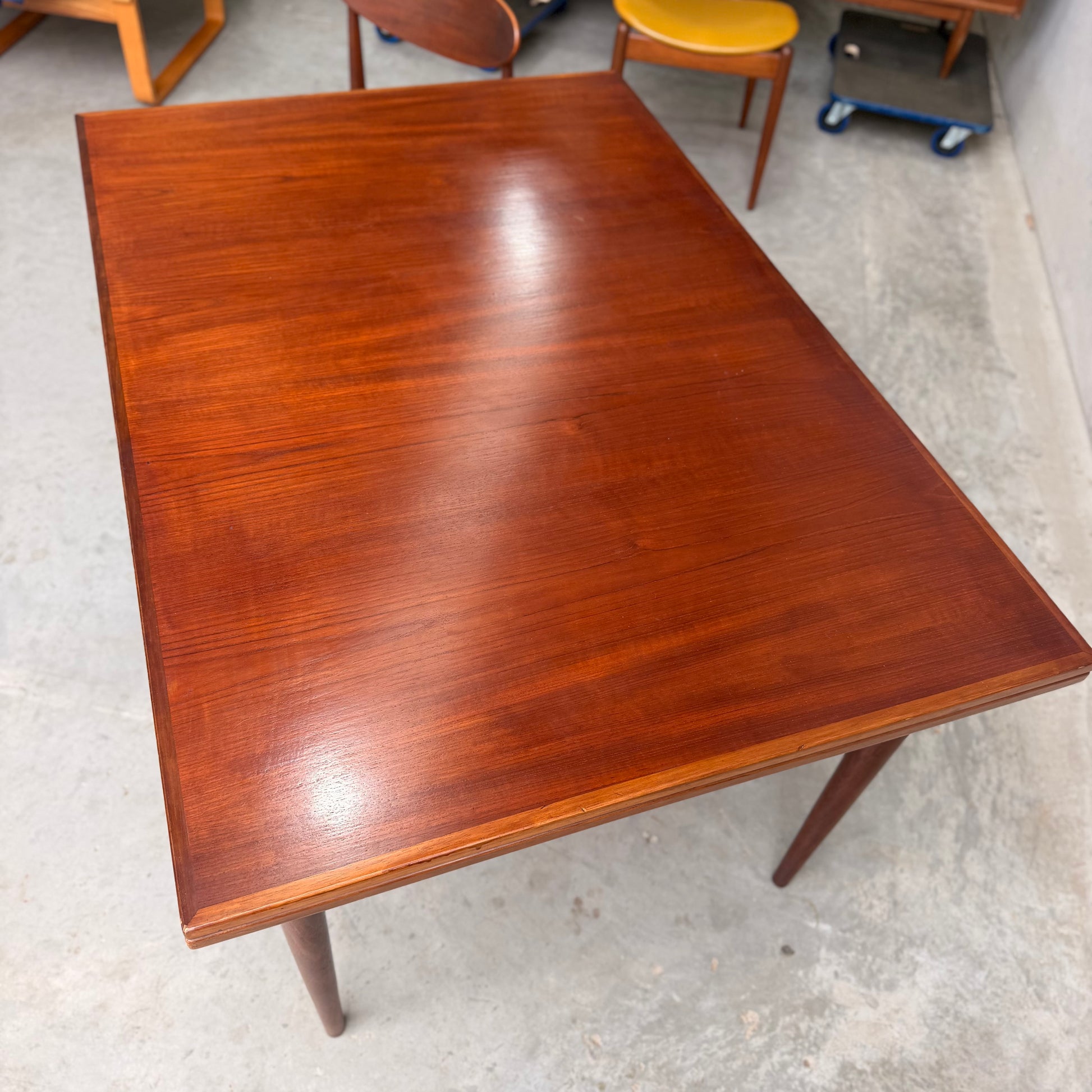 A Vintage Mid-Century Parker 109 Nordic Extension Dining Table (Revive) with a polished surface and tapered legs sits on a concrete floor, surrounded by other mid-century modern chairs and tables in the background.