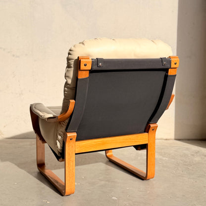 The Tessa Austin Blonde Leather Armchair (Revive), featuring a light wood frame and beige cushioned leather, sits on a concrete floor in bright sunlight, casting a shadow against a plain light-colored wall.