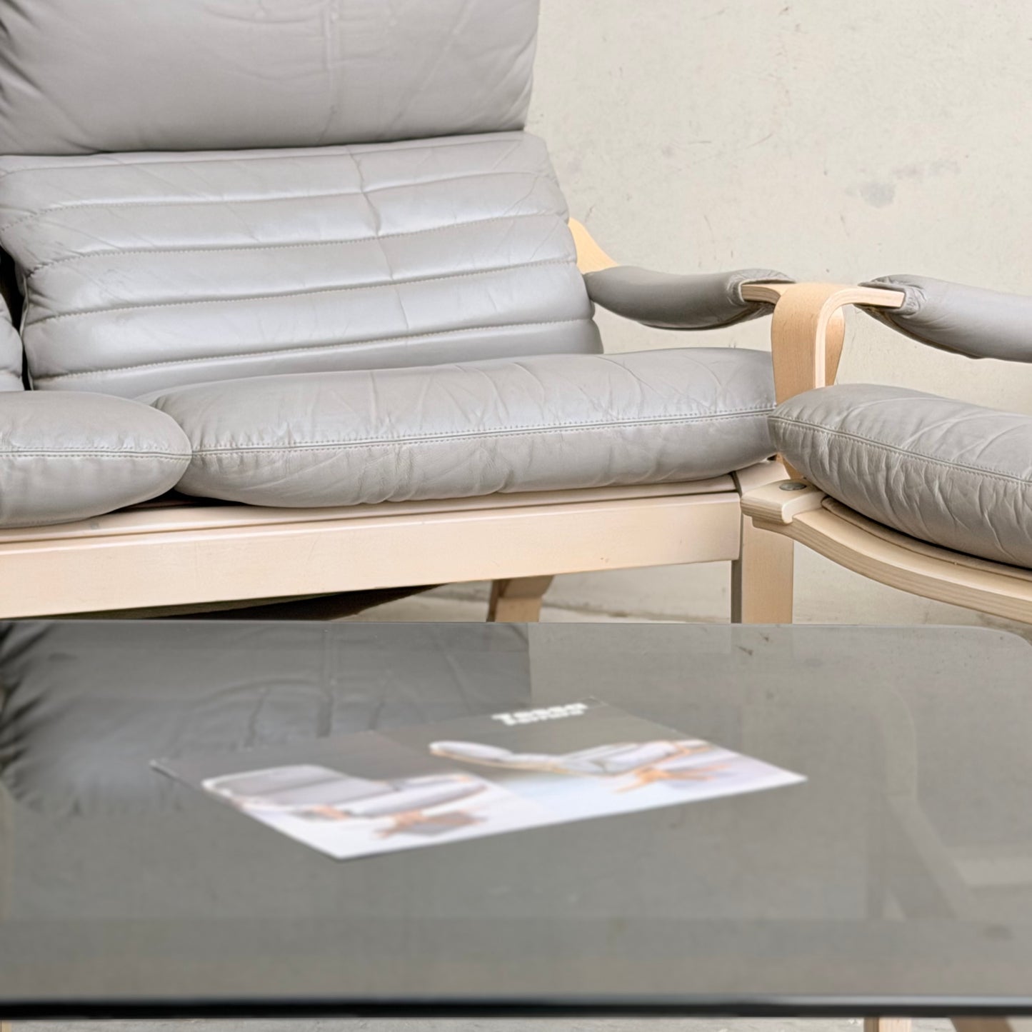A close-up of a light gray cushioned armchair with wooden arms and a matching seat from the Tessa Ferrara Lounge Suite – Complete 5-Piece Set (Revive), paired with a glass coffee table and blurred magazine, adds mid-century modern style against a plain light wall.