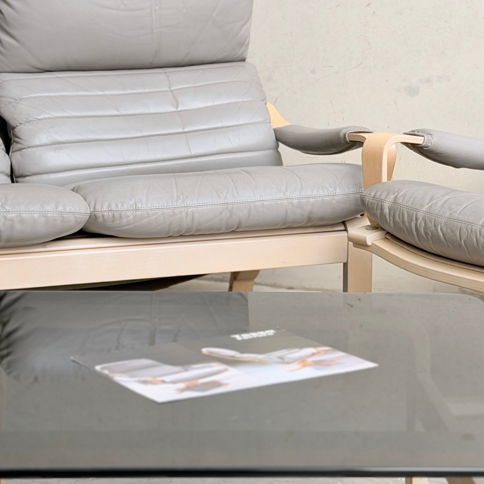 A close-up of a light gray cushioned armchair with wooden arms and a matching seat from the Tessa Ferrara Lounge Suite – Complete 5-Piece Set (Revive), paired with a glass coffee table and blurred magazine, adds mid-century modern style against a plain light wall.