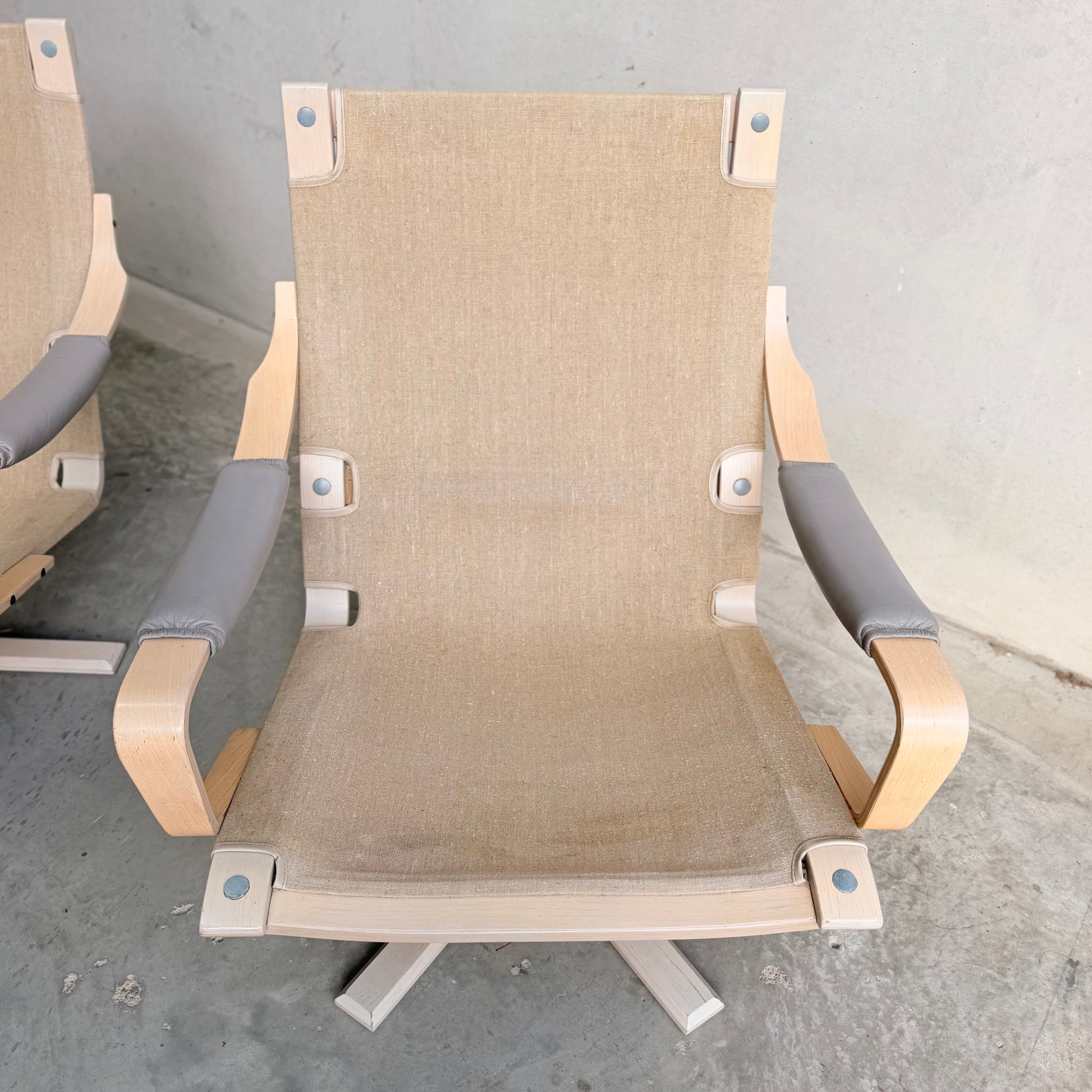 A pair of Tessa Ferrara Swivel Armchairs (Revive) with light wood frames, beige fabric seats and backs, and gray padded armrests sit on a concrete floor against a plain gray wall, highlighting mid-century Australian design.