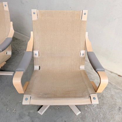 A pair of Tessa Ferrara Swivel Armchairs (Revive) with light wood frames, beige fabric seats and backs, and gray padded armrests sit on a concrete floor against a plain gray wall, highlighting mid-century Australian design.