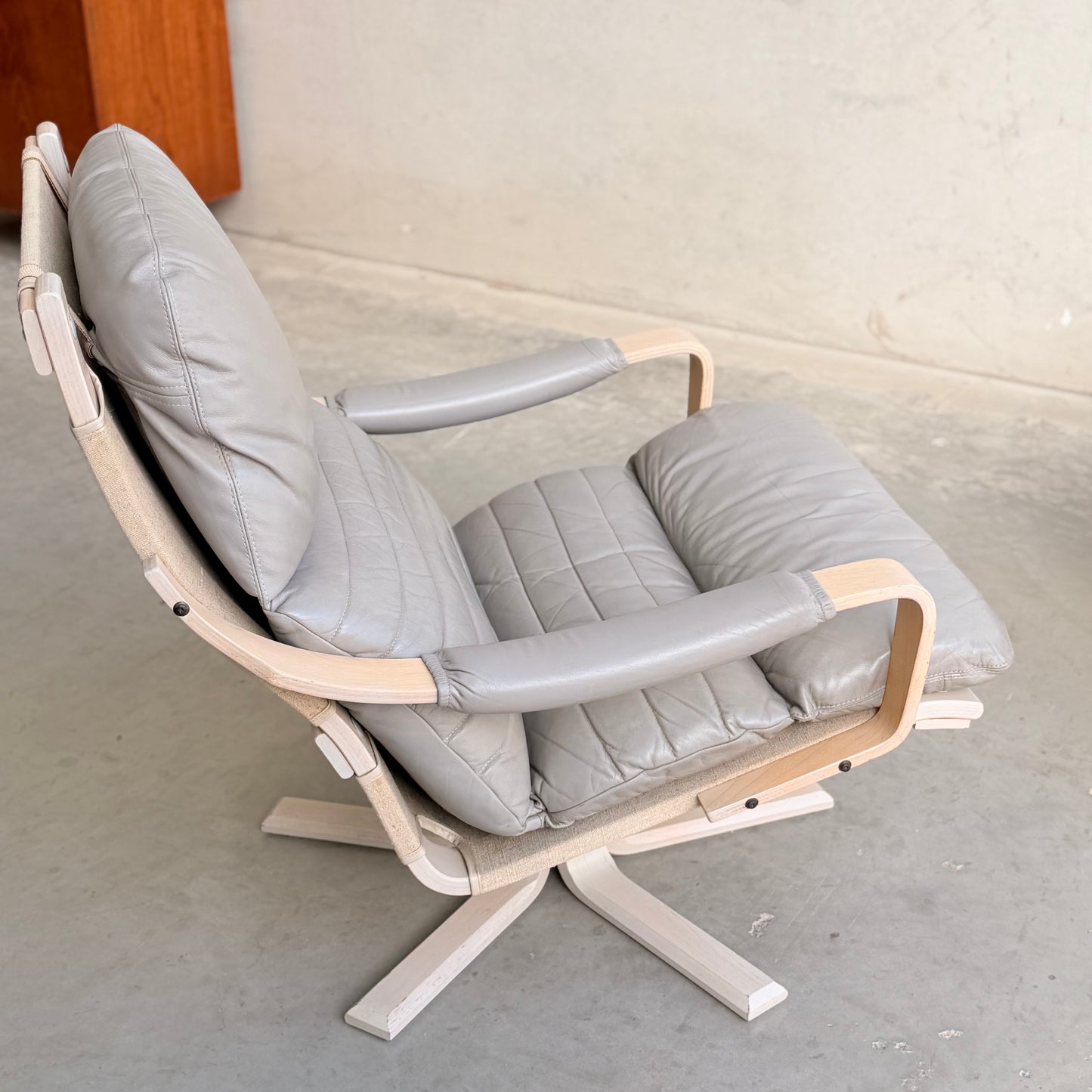 A pair of Tessa Ferrara Swivel Armchairs (Revive) featuring light wood frames and padded light gray leather cushions, showcase iconic mid-century Australian design, set on a concrete floor against a plain wall.