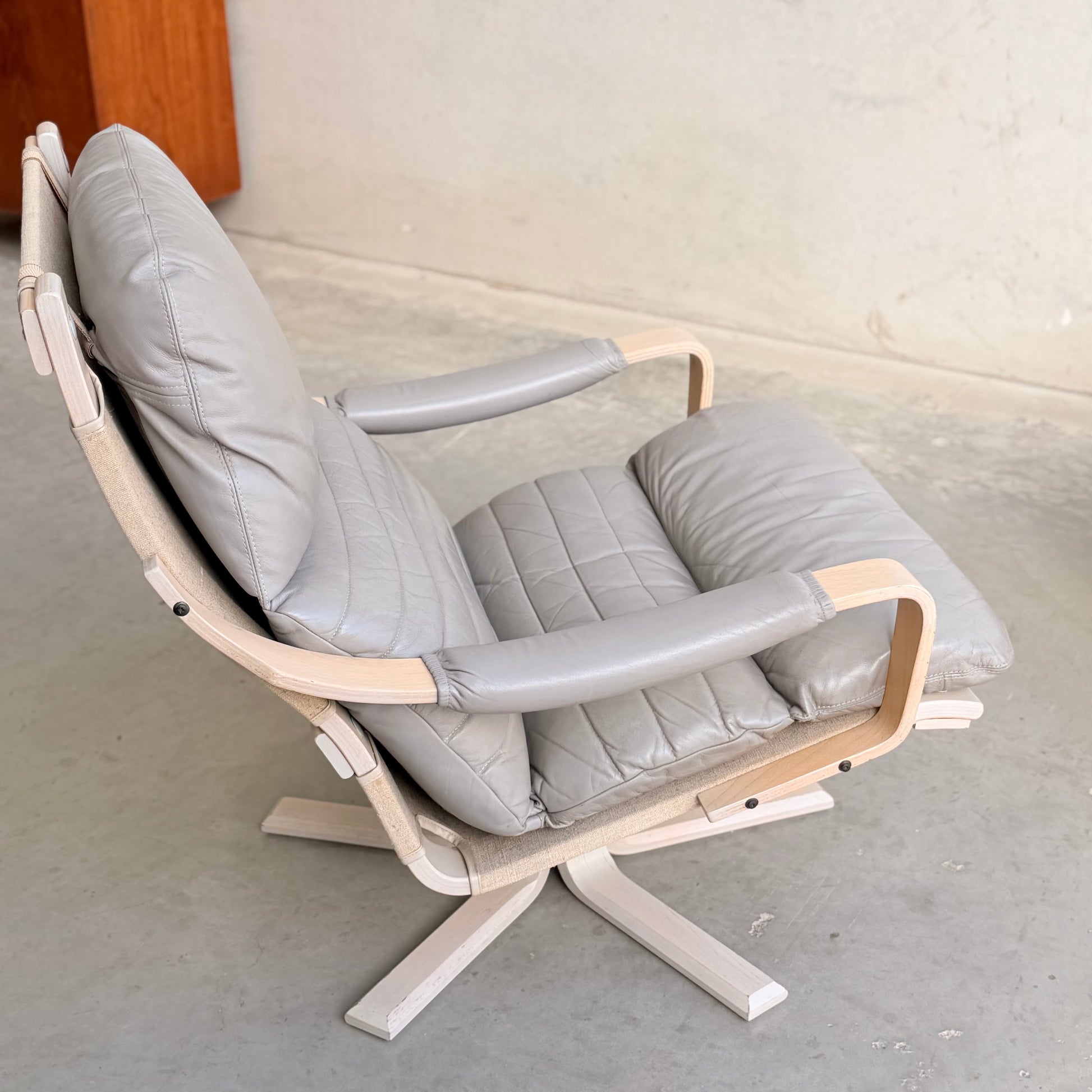 A pair of Tessa Ferrara Swivel Armchairs (Revive) featuring light wood frames and padded light gray leather cushions, showcase iconic mid-century Australian design, set on a concrete floor against a plain wall.