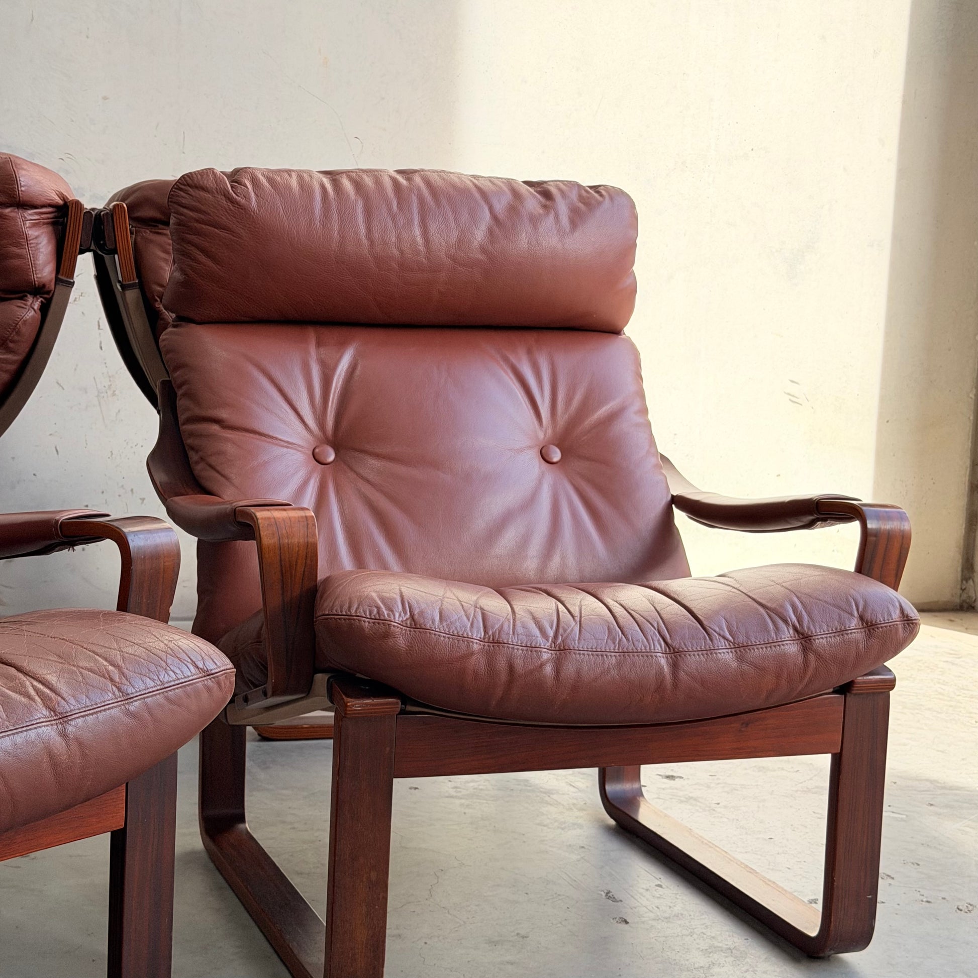 A brown leather armchair with cushioned seat, backrest, and headrest rests on a wooden frame in a sunlit room. This mid-century modern piece is part of the Tessa T8 Lounge Suite modular two-seater sofa & armchairs (Revive).