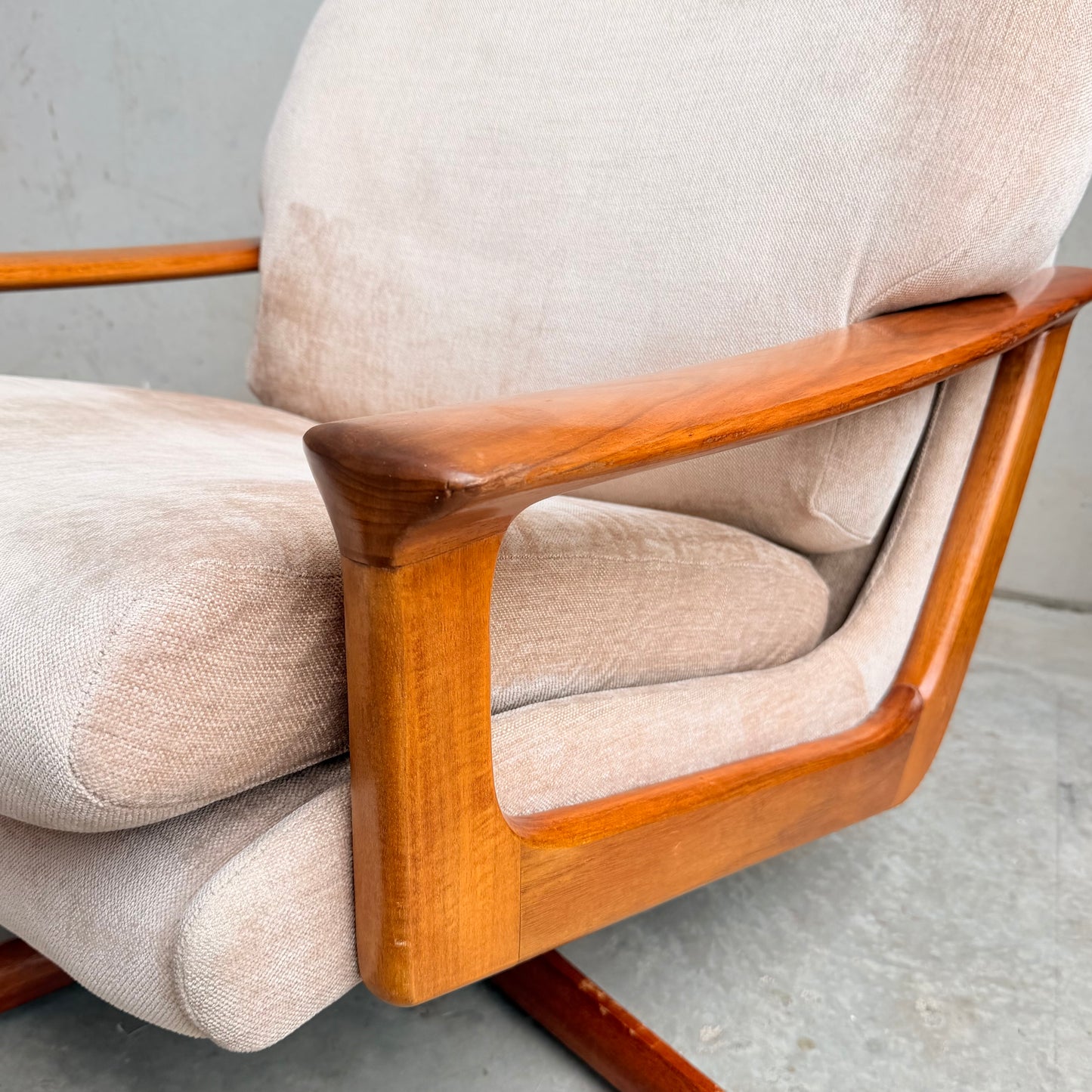 Close-up of the Vintage Mid Century Tessa T21 Swivel Chair (Revive), featuring cream velvet upholstery and smooth curved wooden armrests, set on a concrete floor against a plain grey wall.