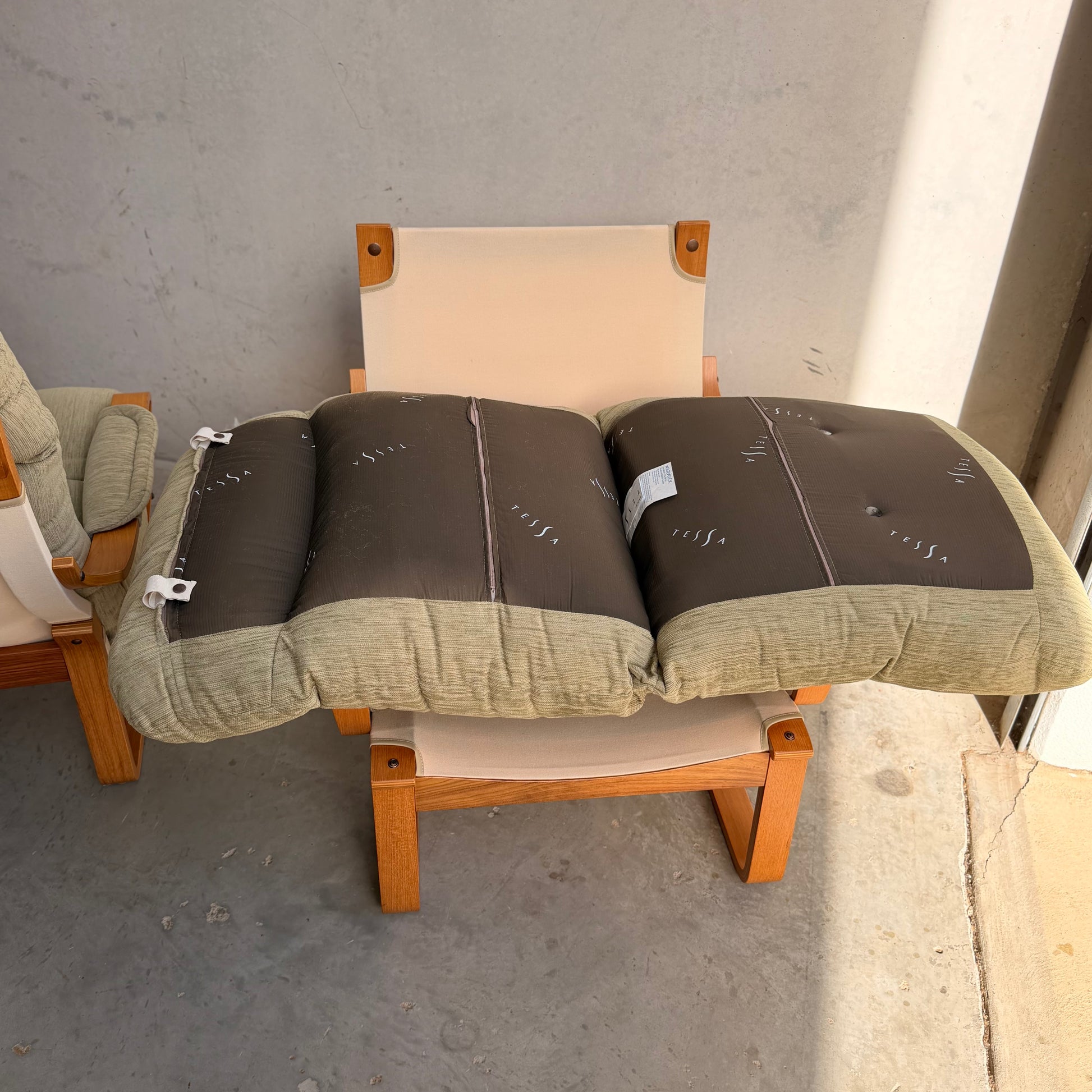 A Pair of Tessa Monaco Arm Chairs - Sage Green Corduroy Like New (Revive) feature a light wood frame and beige seat, accented with two sage corduroy cushions. The mid-century style chairs are placed on a gray concrete floor near a wall.
