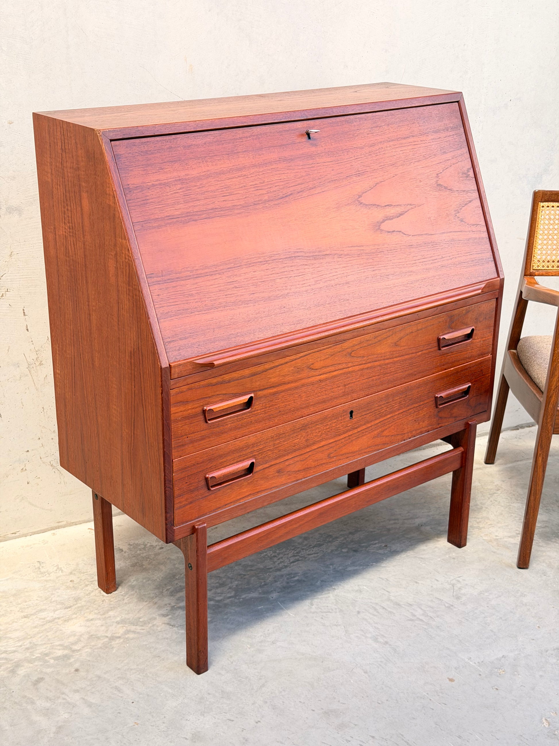 Mid-century modern Danish Arne Wahl Iversen Model 68 teak secretary desk by Vinde Møbelfabrik, featuring three drawers and a slanted drop-front, paired with a cane-back wooden chair against a light wall and concrete floor.