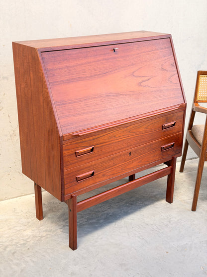 Mid-century modern Danish Arne Wahl Iversen Model 68 teak secretary desk by Vinde Møbelfabrik, featuring three drawers and a slanted drop-front, paired with a cane-back wooden chair against a light wall and concrete floor.