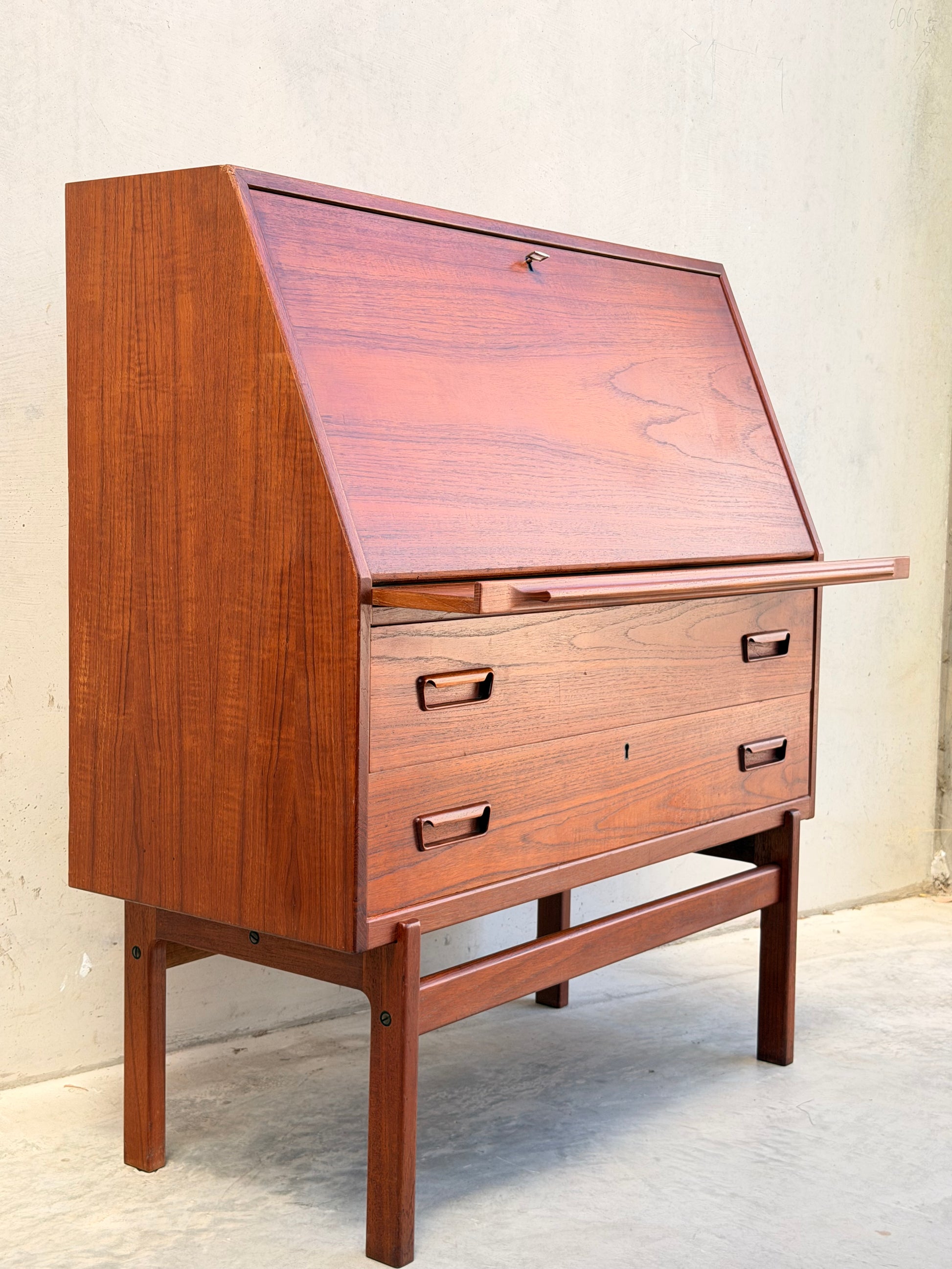 A Mid Century Modern Danish Arne Wahl Iversen Model 68 Teak Secretary Desk by Vinde Møbelfabrik, featuring a slanted drop-down writing surface, upper small drawers, and lower large drawers, stands on four legs against a plain light wall.