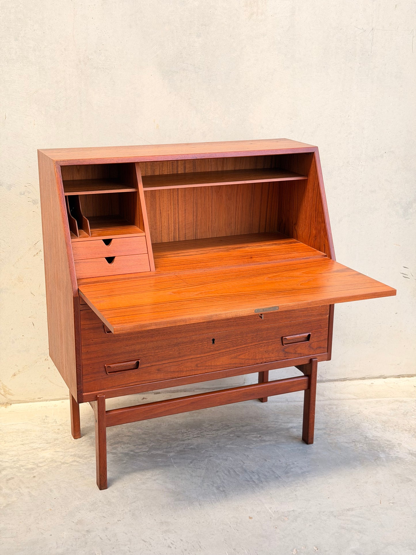 A Mid Century Modern Danish Arne Wahl Iversen Model 68 Teak Secretary Desk by Vinde Møbelfabrik, featuring open cubbies, small drawers, and a drop-down writing surface, sits against a plain light-colored wall and concrete floor.