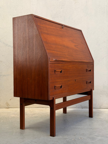 A Mid Century Modern Danish Arne Wahl Iversen Model 68 Teak Secretary Desk by Vinde Møbelfabrik, featuring a slanted front and two lower drawers, stands on four legs against a light wall and concrete floor.