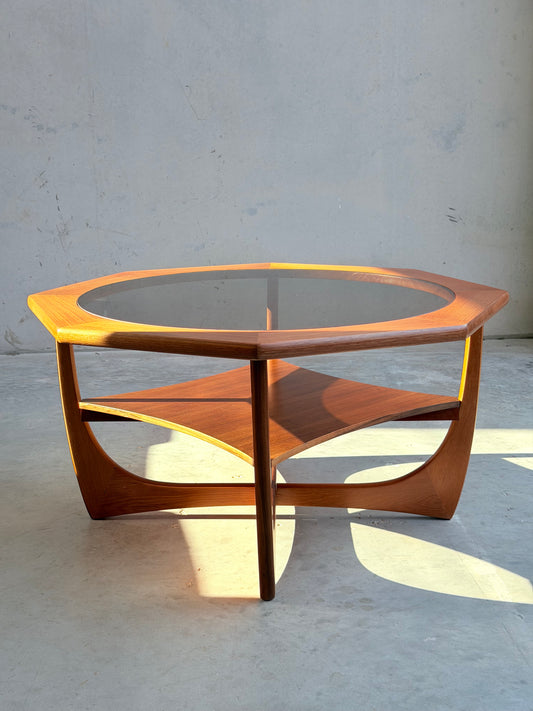 An octagonal mid-century Kalmar Furniture coffee table with a smoked glass top and hardwood magazine rack stands against a plain concrete wall, sunlight casting shadows on the floor.