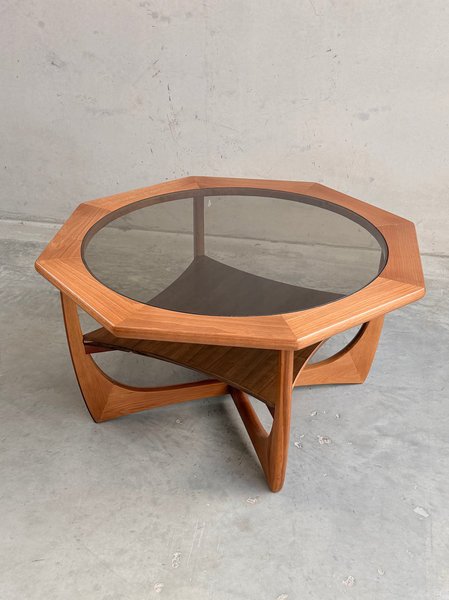 An Octagonal Mid Century Coffee Table by Kalmar Furniture, made of hardwood with smoked glass and a magazine rack, stands on a gray concrete floor against a plain light wall.
