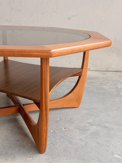 An Octagonal Mid Century Coffee Table by Kalmar Furniture, featuring a smoked glass top and hardwood magazine rack, stands on a concrete floor against a plain light wall—highlighting classic Australian design.