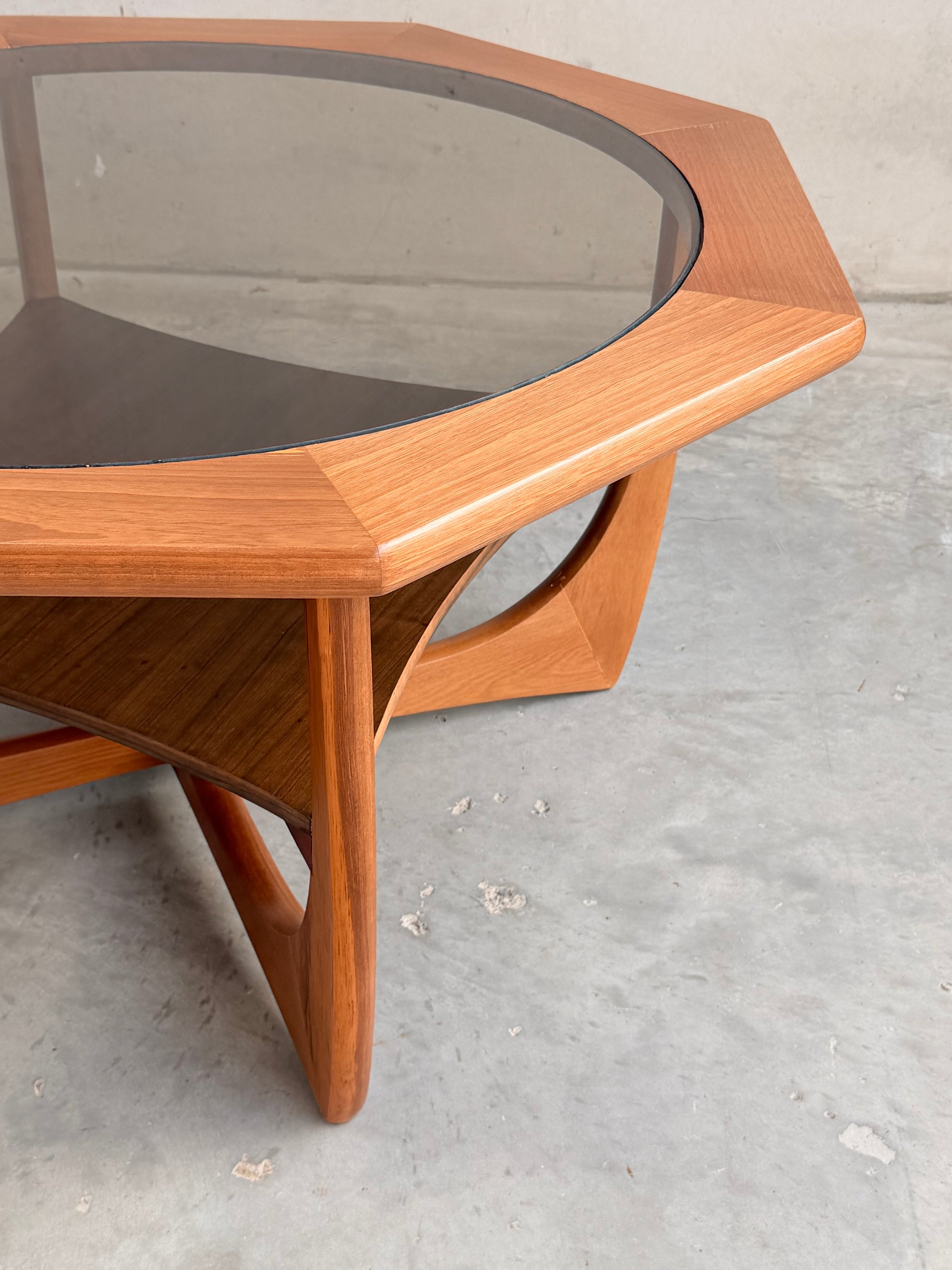 The Octagonal Mid Century Coffee Table by Kalmar Furniture features hardwood construction, a smoked glass top, and an integrated magazine rack (Revive), highlighting Australian design on a concrete floor against a plain wall.