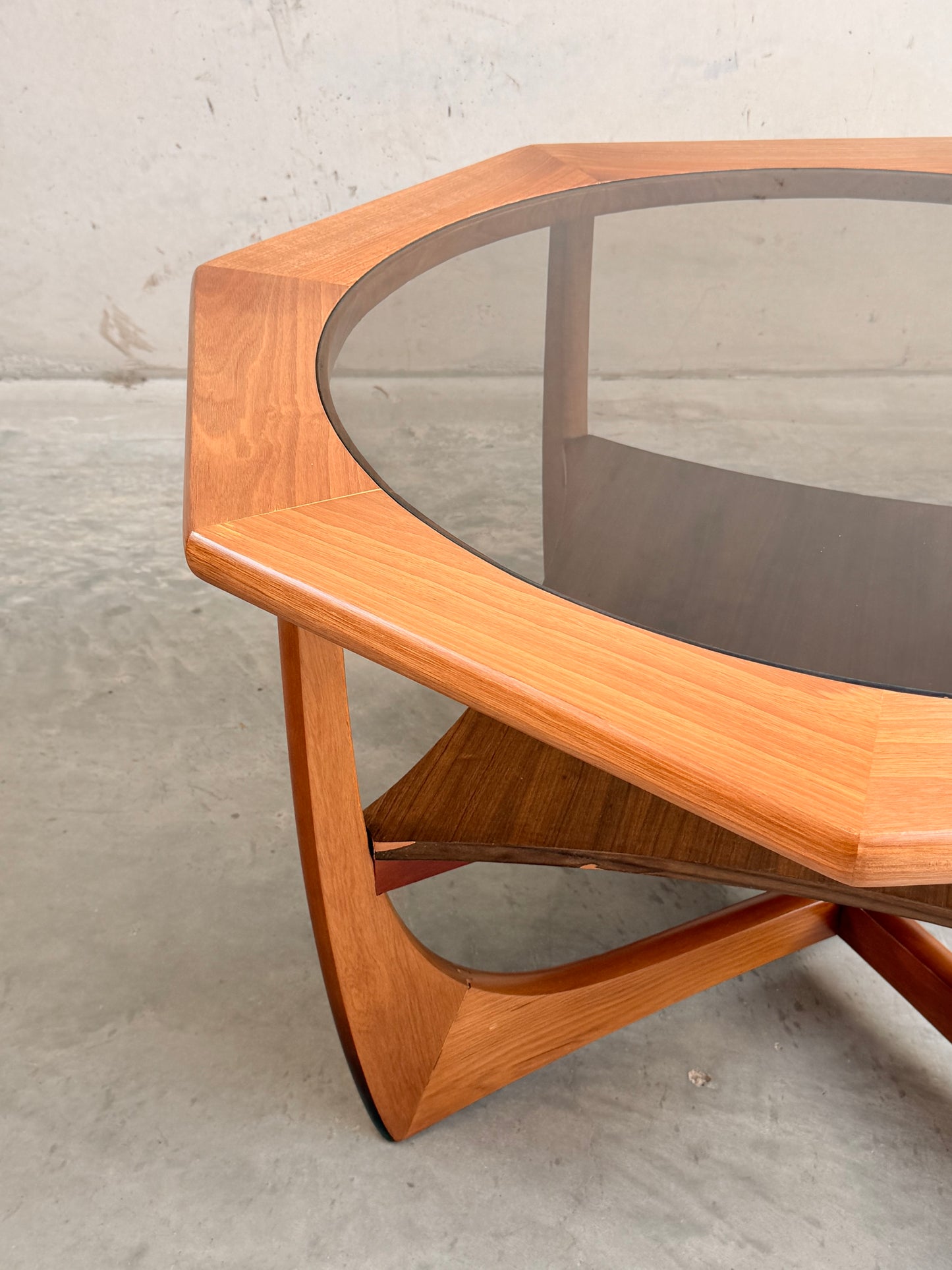 The Octagonal Mid Century Coffee Table by Kalmar Furniture features a hardwood frame, smoked glass top, and lower magazine rack, exemplifying mid-century style—shown on a concrete floor against a light-colored wall.