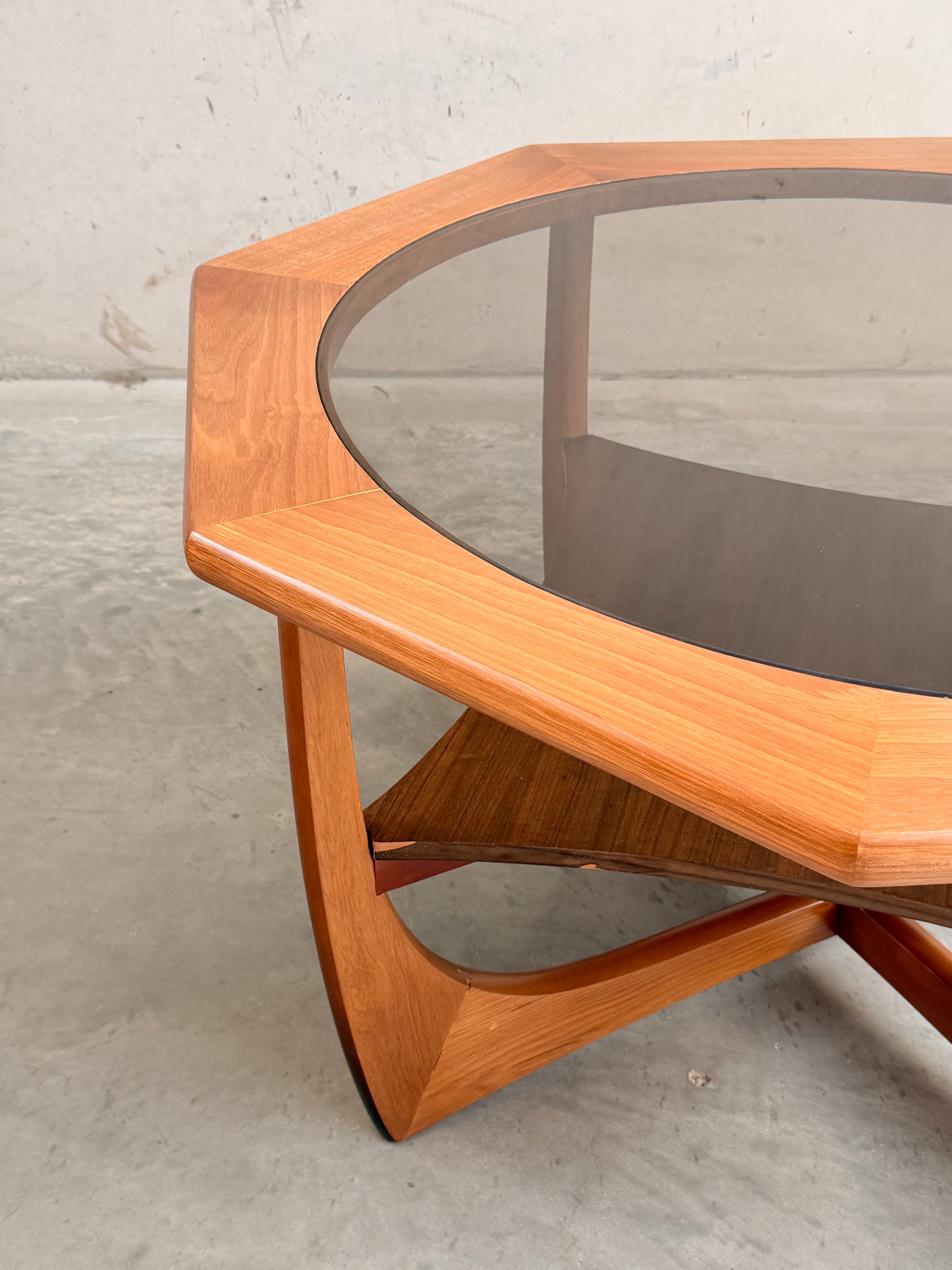 The Octagonal Mid Century Coffee Table by Kalmar Furniture features a hardwood frame, smoked glass top, and lower magazine rack, exemplifying mid-century style—shown on a concrete floor against a light-colored wall.