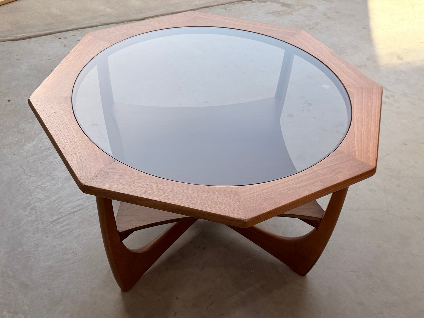The Octagonal Mid Century Coffee Table by Kalmar Furniture features hardwood construction, a smoked glass top, and a built-in magazine rack, styled in Australian design and placed on a smooth concrete floor.