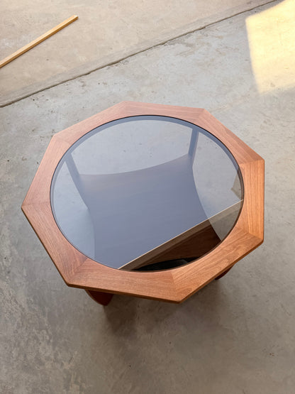 An Octagonal Mid Century Coffee Table by Kalmar Furniture, featuring a hardwood base, smoked glass top, and magazine rack (Revive), sits on a concrete floor casting a faint shadow, evoking timeless Australian design. A wooden stick lies in the background.