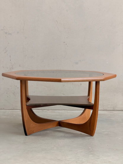 An Octagonal Mid Century Coffee Table by Kalmar Furniture, crafted from hardwood with smoked glass and a built-in magazine rack, stands out as an elegant Australian design icon against a plain, light-colored wall.
