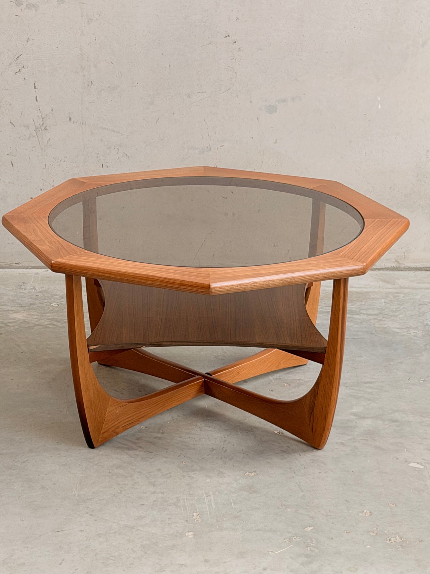 The Octagonal Mid Century Coffee Table by Kalmar Furniture features hardwood construction, a smoked glass top, and a magazine rack, embodying vintage style against a plain, light-colored concrete backdrop.