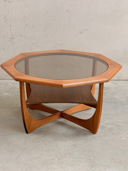 The Octagonal Mid Century Coffee Table by Kalmar Furniture features hardwood construction, a smoked glass top, and a magazine rack, embodying vintage style against a plain, light-colored concrete backdrop.