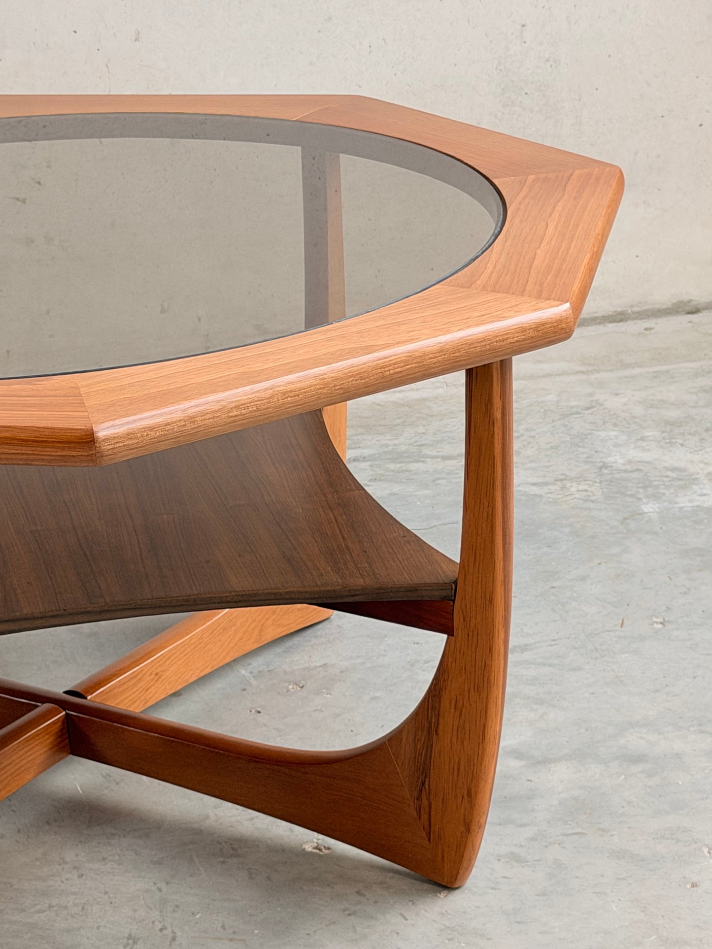 A close-up of the Octagonal Mid Century Coffee Table by Kalmar Furniture, featuring smoked glass, hardwood frame, geometric angles, and a curved magazine rack shelf below, displayed on a smooth concrete floor.