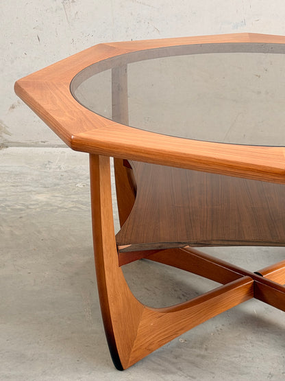An octagonal mid-century coffee table by Kalmar Furniture, featuring a smoked glass top, hardwood frame, and magazine rack (Revive), is set against a plain light concrete background.