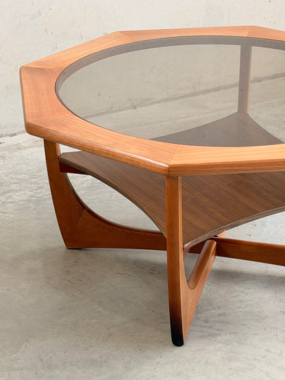 An Octagonal Mid Century Coffee Table by Kalmar Furniture features a hardwood frame, smoked glass top, and magazine rack. With classic Australian design and angled legs, it brings smooth mid-century style to any space. Shown on concrete against a plain wall.
