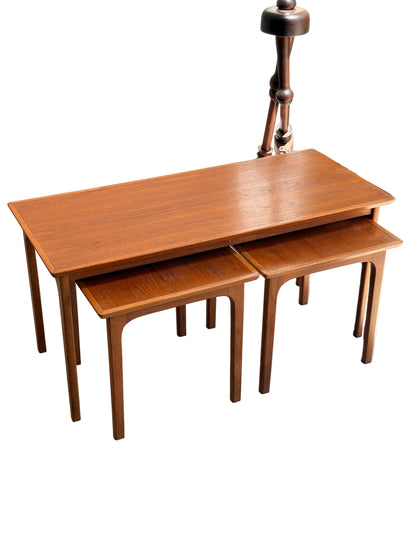 A set of Classic Australian-Made Noblett Mid-Century Teak Nesting Tables – Circa 1970s (Revive), with two smaller tables partially tucked under the larger one, is displayed on a light concrete floor near a wall and window.