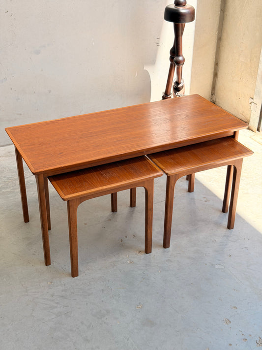 A set of Classic Australian-Made Noblett Mid-Century Teak Nesting Tables – Circa 1970s (Revive), with two smaller tables partially tucked under the larger one, is displayed on a light concrete floor near a wall and window.