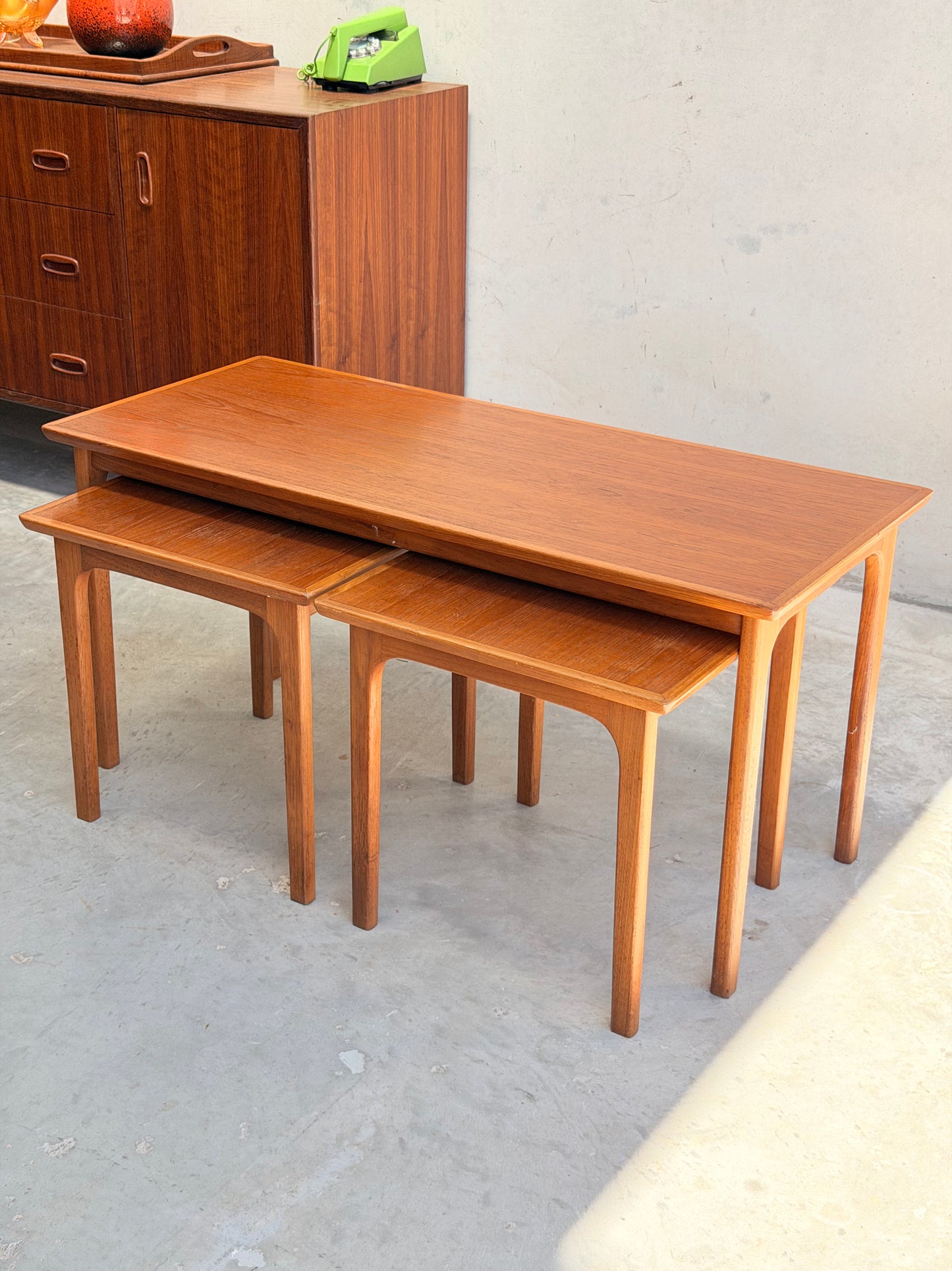 A set of Classic Australian-Made Noblett Mid-Century Teak Nesting Tables – Circa 1970s (Revive), with tapered legs and Scandinavian design, displayed staggered on a light floor next to a matching cabinet and green rotary phone.