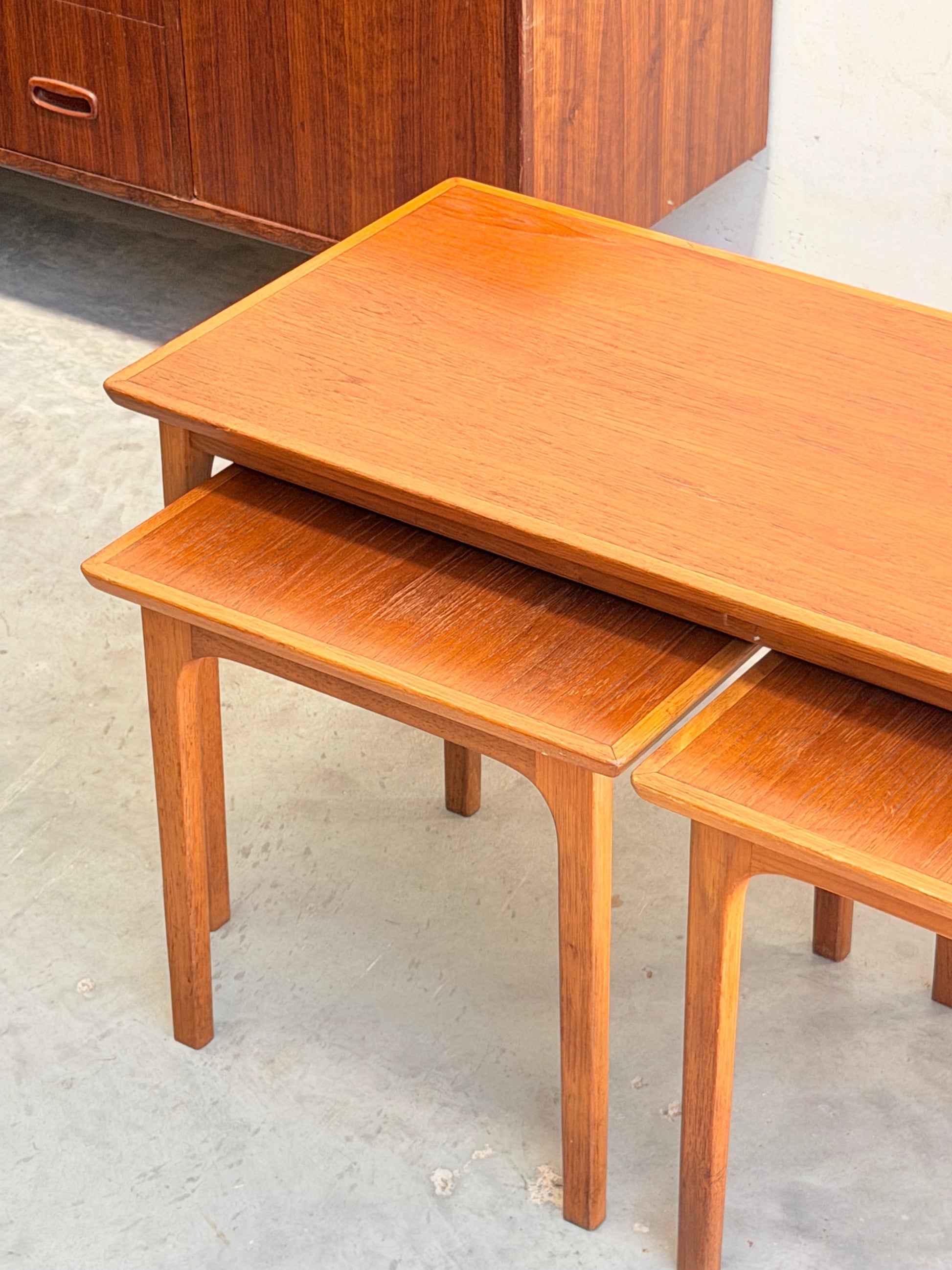 Two Classic Australian-Made Noblett Mid-Century Teak Nesting Tables (circa 1970s, Revive) with rectangular tops and straight legs sit on a gray concrete floor; another piece of mid-century teak furniture appears in the background.