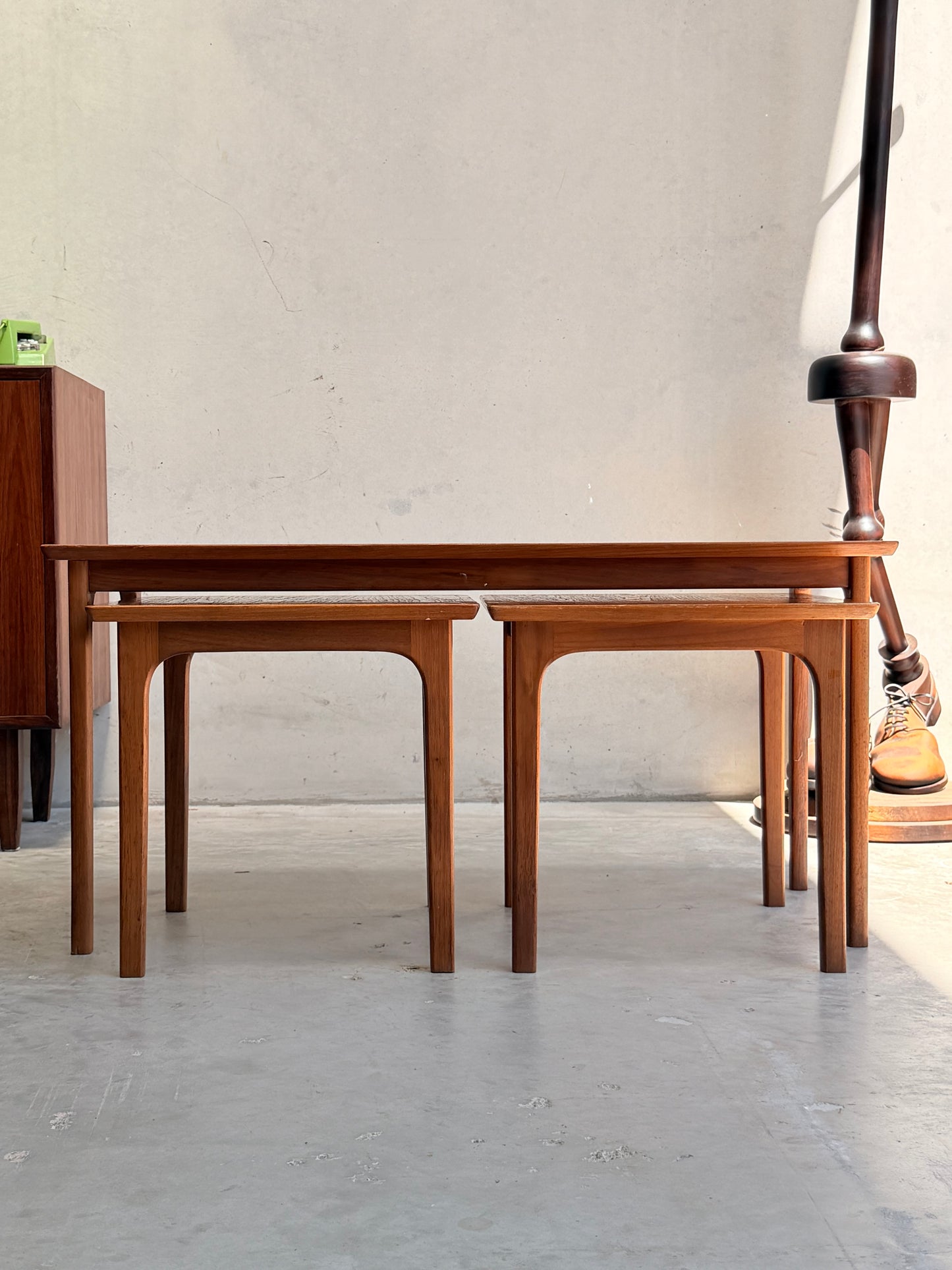 Two Classic Australian-Made Noblett Mid-Century Teak Nesting Tables (circa 1970s, Revive) sit side by side on a light floor against a beige wall, sunlight casting gentle shadows. A floor lamp with a round base is partially visible on the right.