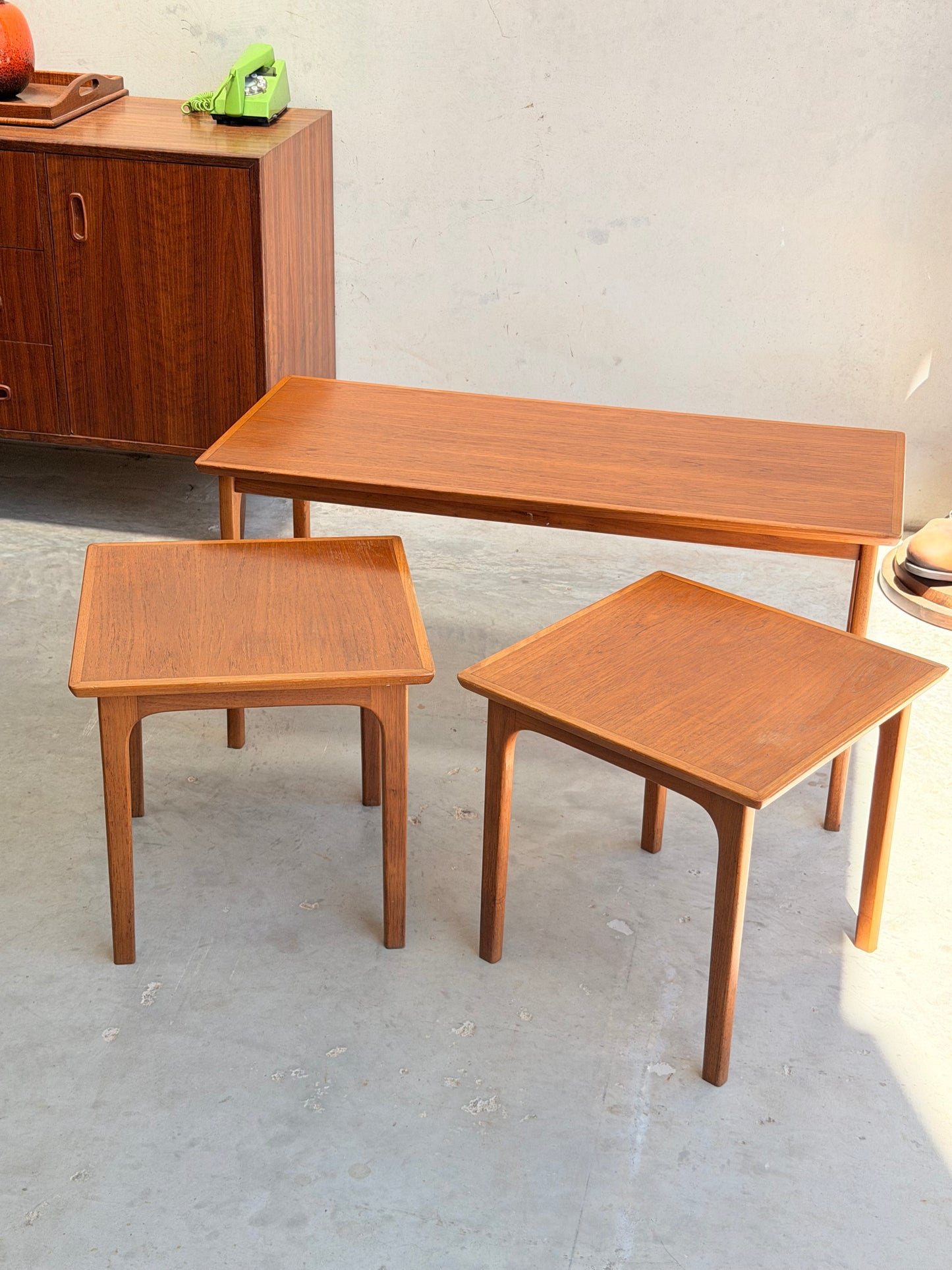 Three Classic Australian-Made Noblett Mid-Century Teak Nesting Tables – Circa 1970s (Revive) are arranged on a light gray floor near a vintage cabinet with a green rotary phone, sunlight emphasizing their elegant Scandinavian-inspired design.