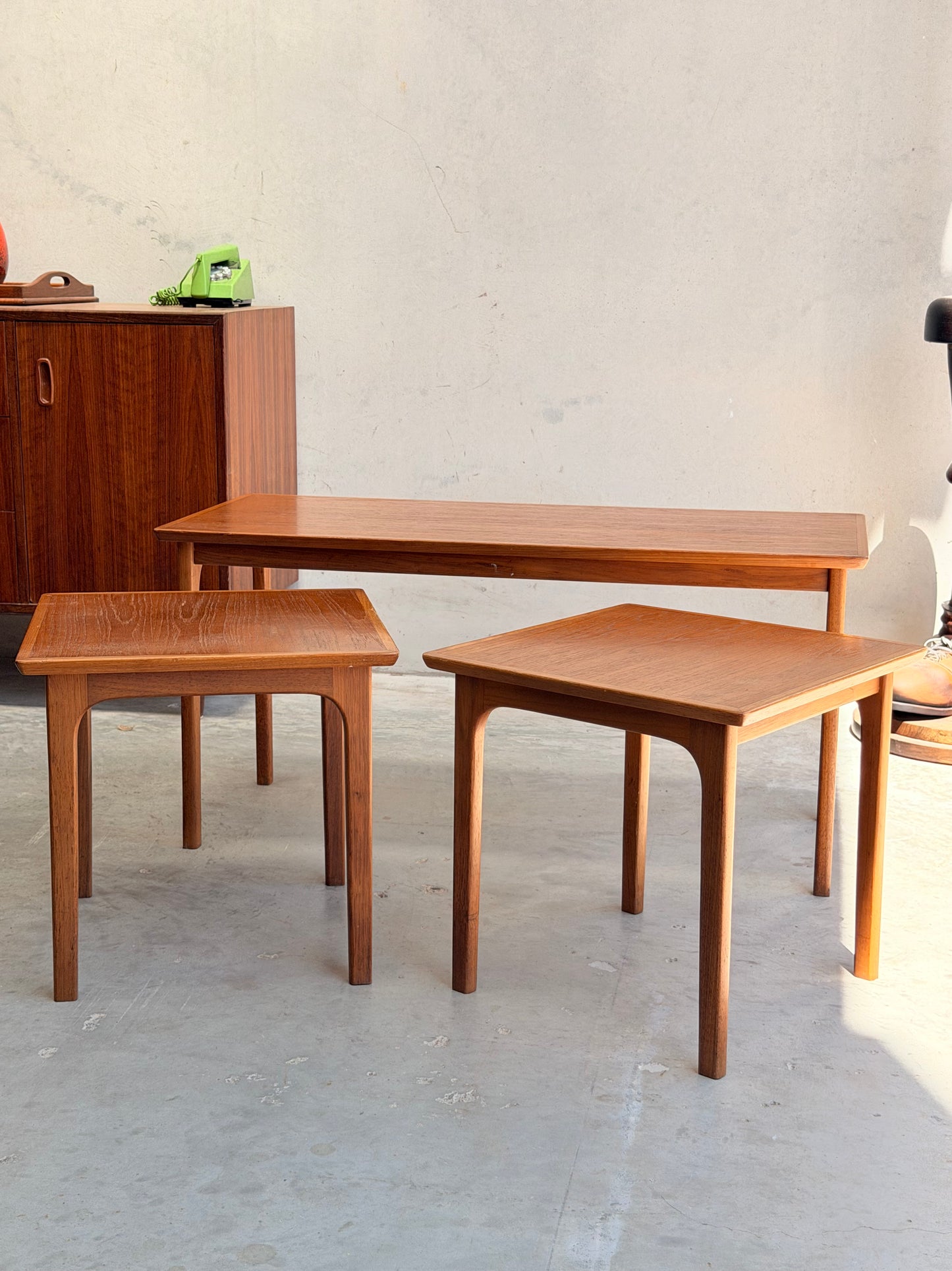 Three Classic Australian-Made Noblett Mid-Century Teak Nesting Tables – Circa 1970s (Revive) of varying heights are arranged on a light gray floor, with a wood cabinet and green rotary phone in the background against a plain beige wall.