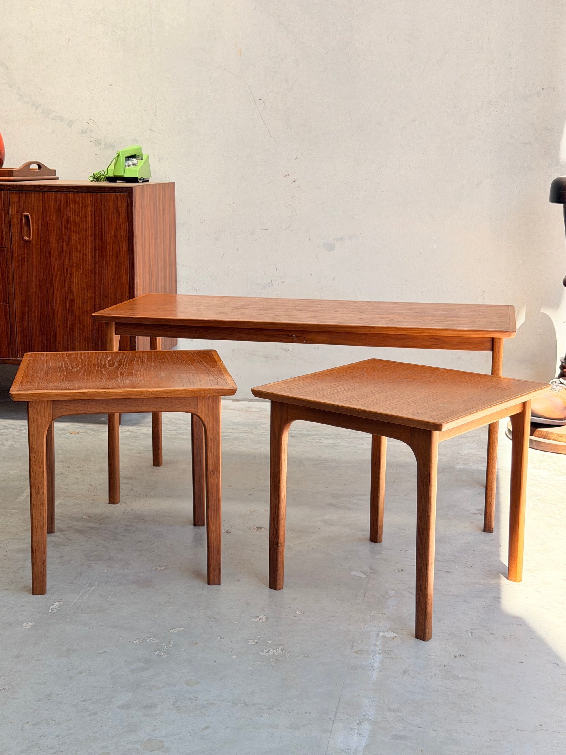 Three Classic Australian-Made Noblett Mid-Century Teak Nesting Tables – Circa 1970s (Revive) of varying heights are arranged on a light gray floor, with a wood cabinet and green rotary phone in the background against a plain beige wall.