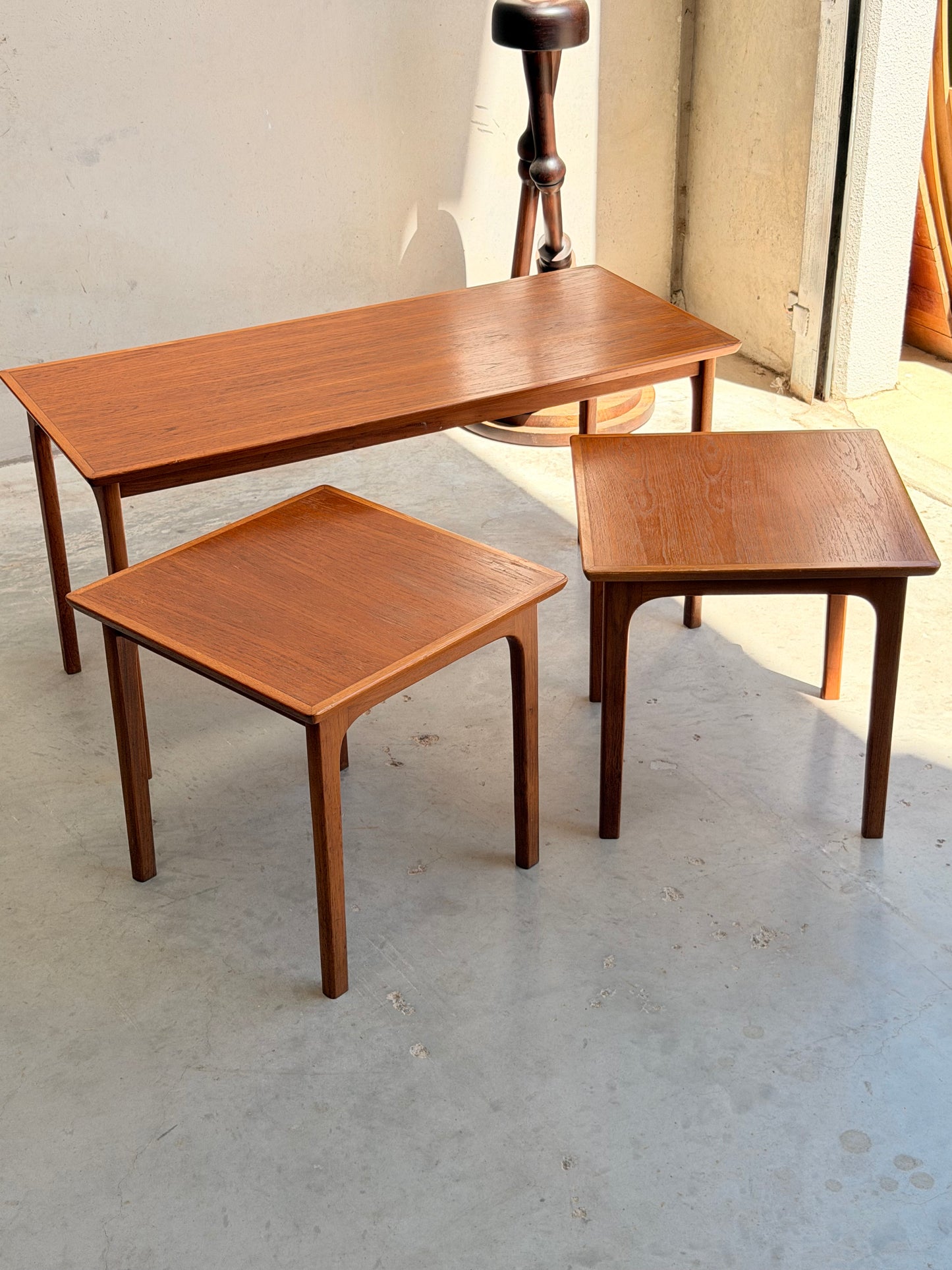 Three Classic Australian-Made Noblett Mid-Century Teak Nesting Tables (circa 1970s, Revive) of different sizes are arranged on a light concrete floor near a plain wall and an open doorway with sunlight streaming in.