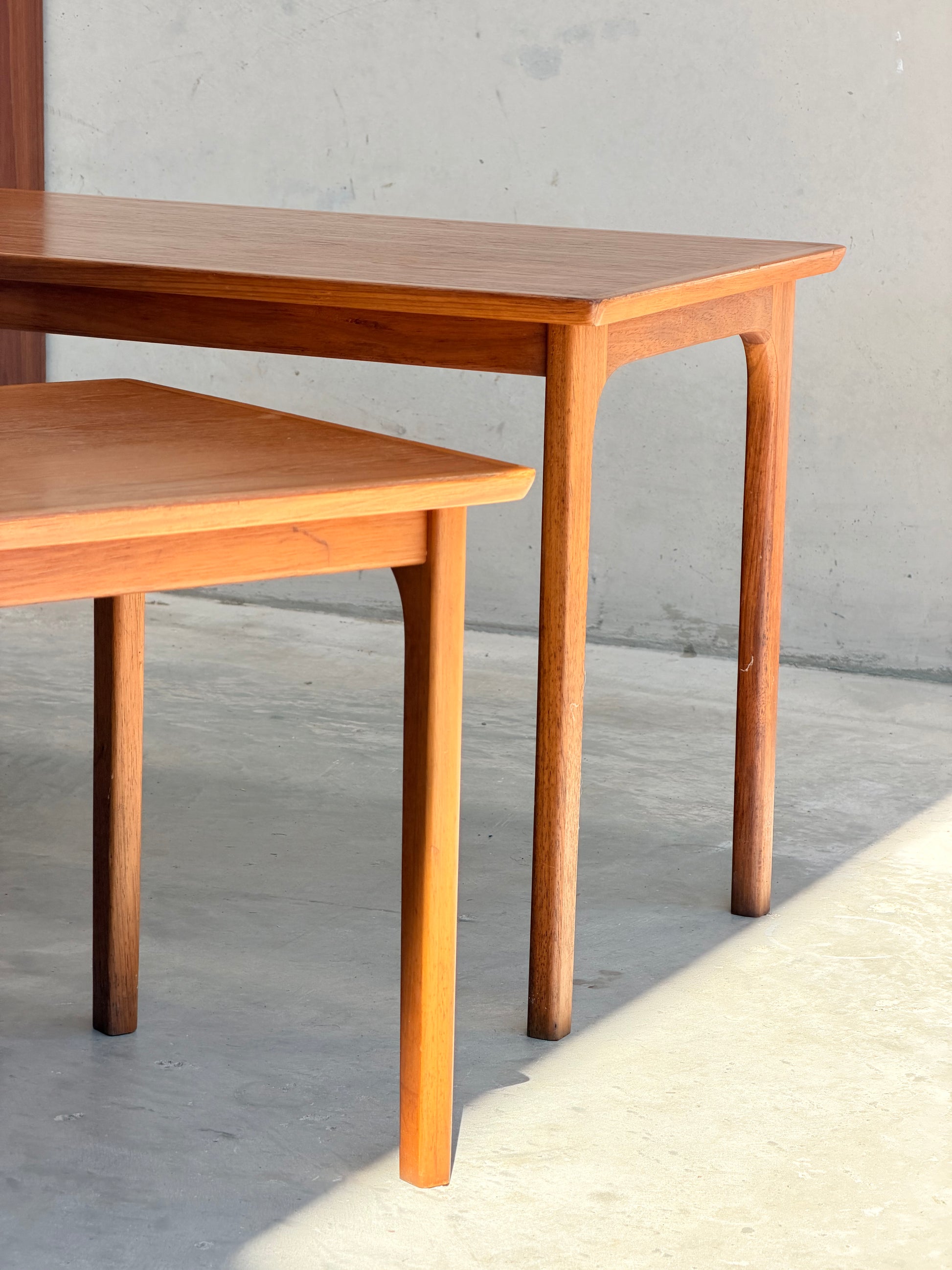 Two Classic Australian-Made Noblett Mid-Century Teak Nesting Tables – Circa 1970s (Revive) highlight timeless Scandinavian design, bathed in sunlight against a pale wall and casting gentle shadows across a concrete floor.