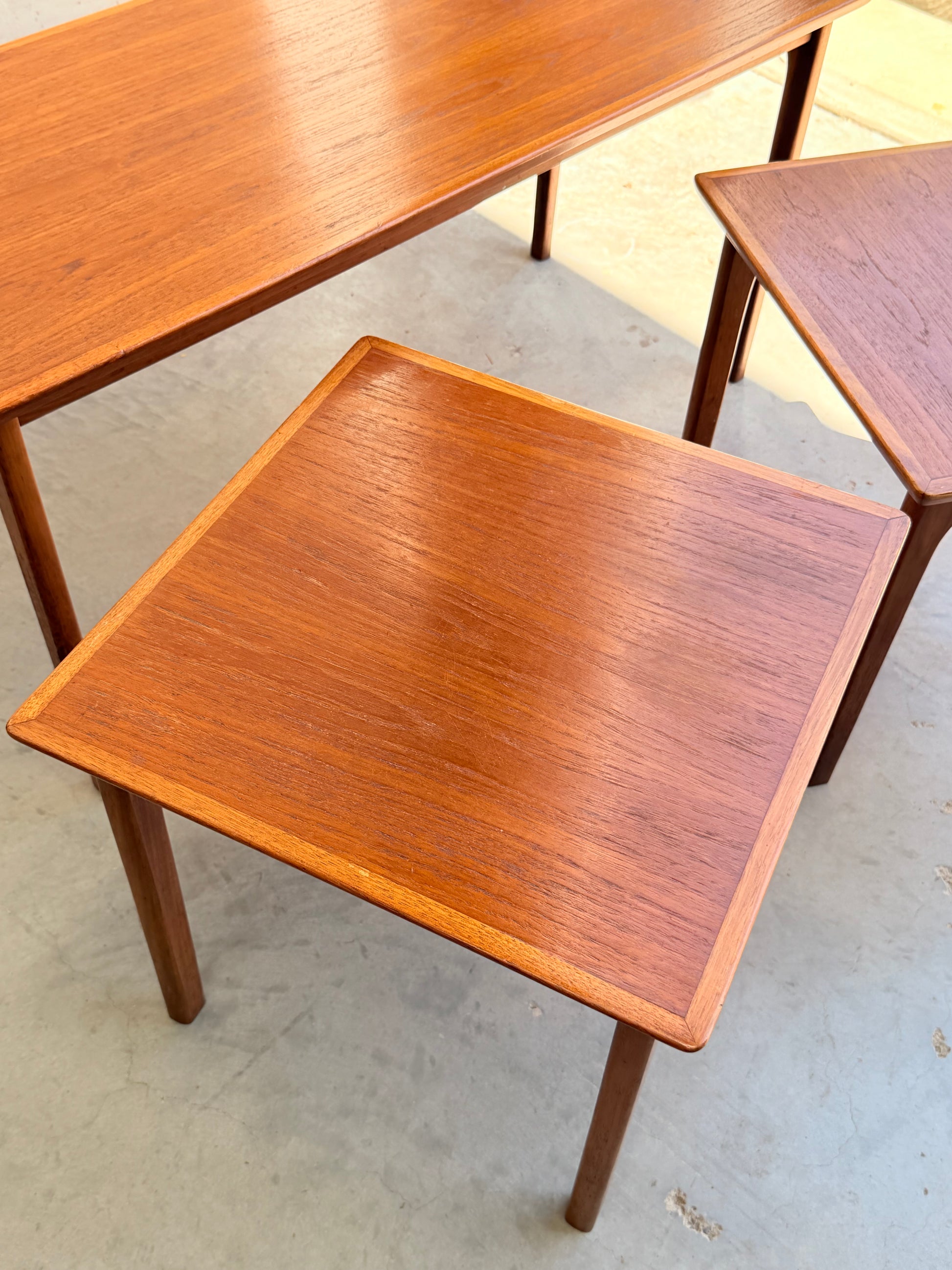 Three Classic Australian-Made Noblett Mid-Century Teak Nesting Tables – Circa 1970s (Revive) with elegant legs and smooth tops are displayed on a light concrete floor, their warm finish echoing iconic Scandinavian design.