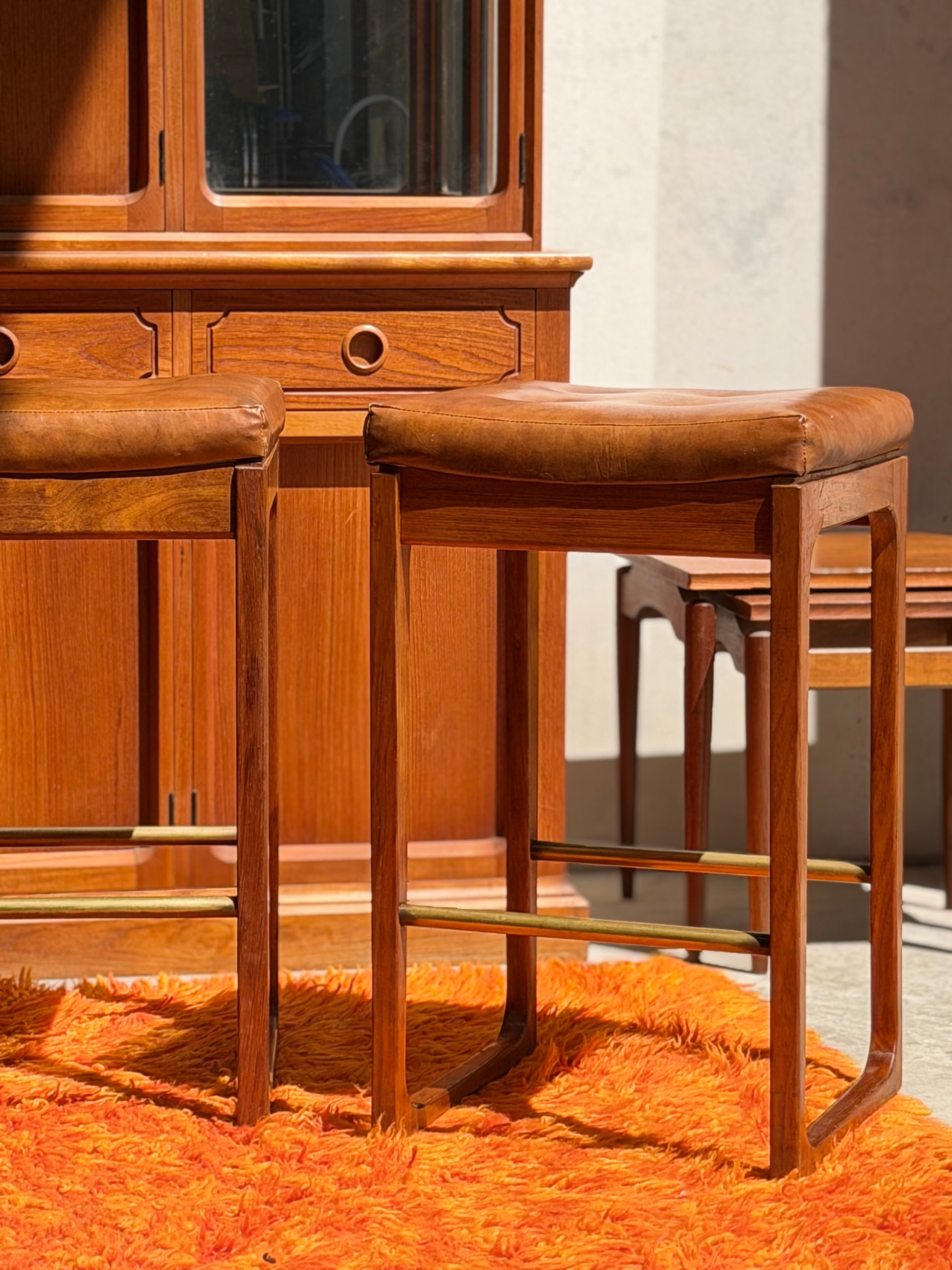 Four Gerald Easden ‘Module’ 1960s Blackwood Bar Stools (Revive) with brown leather seats sit on a bright orange shag rug in sunlight, framed by a wooden cabinet and side tables in the background.
