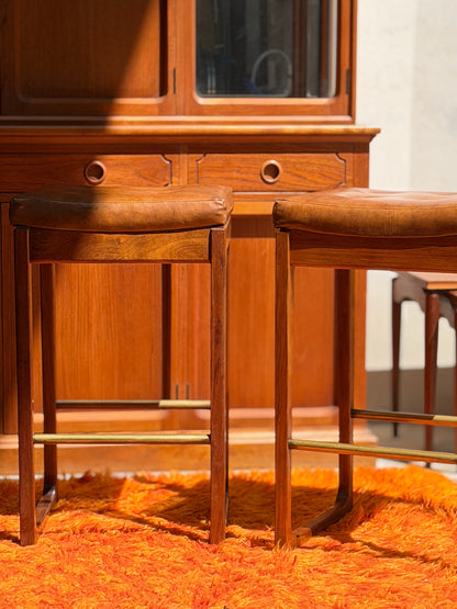 Four Gerald Easden ‘Module’ 1960s Blackwood Bar Stools (Revive) with brown leather seats are arranged on an orange shag rug before a wooden cabinet, sunlight casting shadows across the scene.