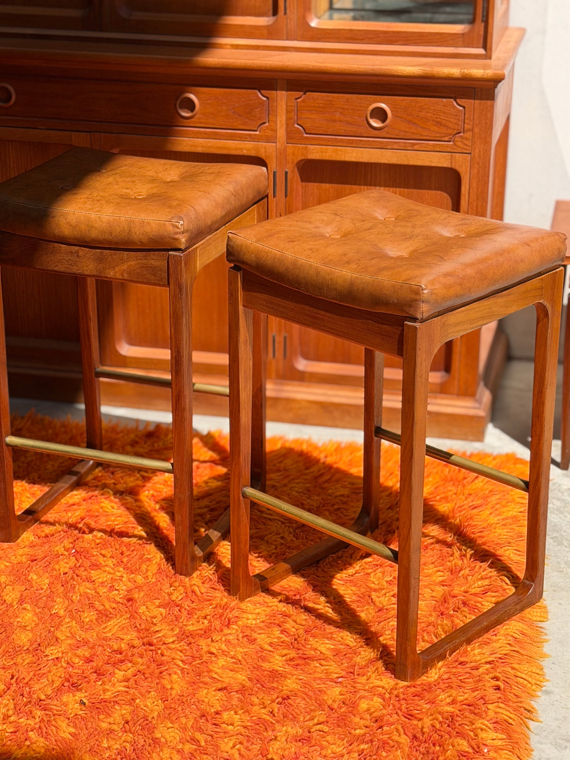 A set of Gerald Easden ‘Module’ 1960s Blackwood Bar Stools (Revive) sits on a shaggy orange rug in front of a wooden cabinet, all bathed in sunlight.