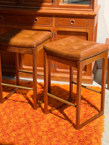 A set of Gerald Easden ‘Module’ 1960s Blackwood Bar Stools (Revive) sits on a shaggy orange rug in front of a wooden cabinet, all bathed in sunlight.