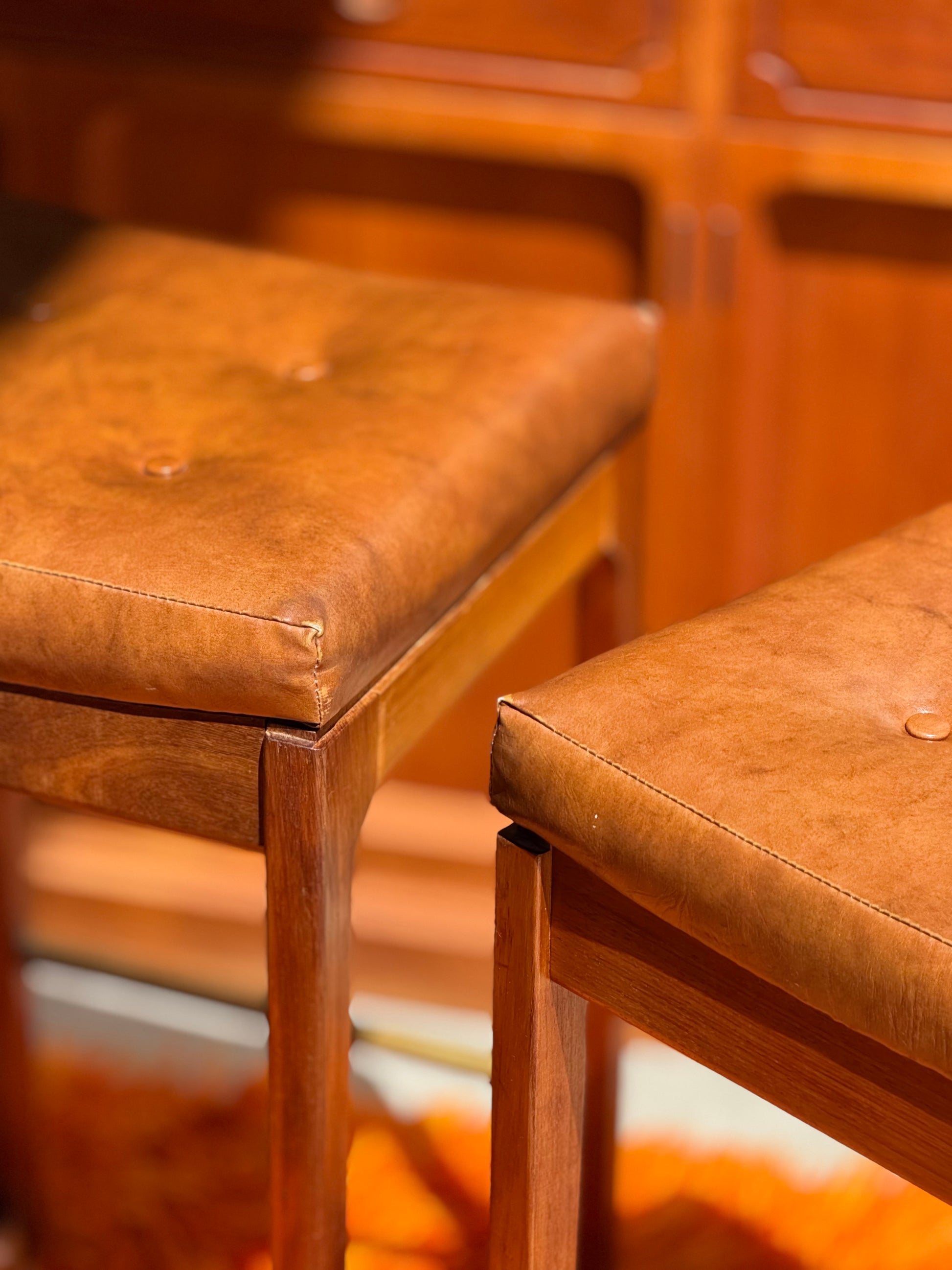 A set of four Gerald Easden ‘Module’ 1960s blackwood bar stools (Revive) with padded brown leather seats, set against matching wood furniture, evokes a warm, vintage mid-century modern ambiance.