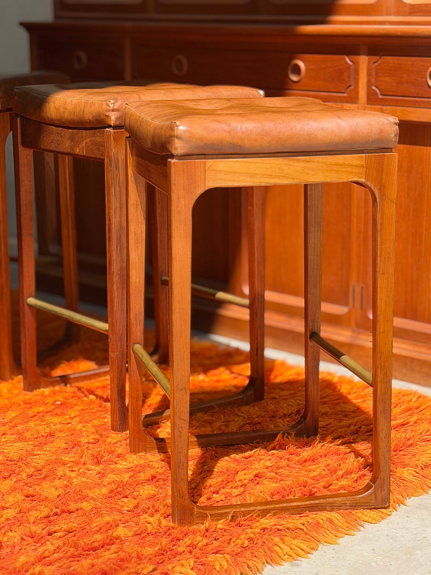 Four Gerald Easden ‘Module’ 1960s Blackwood Bar Stools (Revive) with brown leather seats are arranged on a bright orange shag rug in front of a wooden cabinet, their textures and colors enhanced by warm lighting for a mid-century modern vibe.